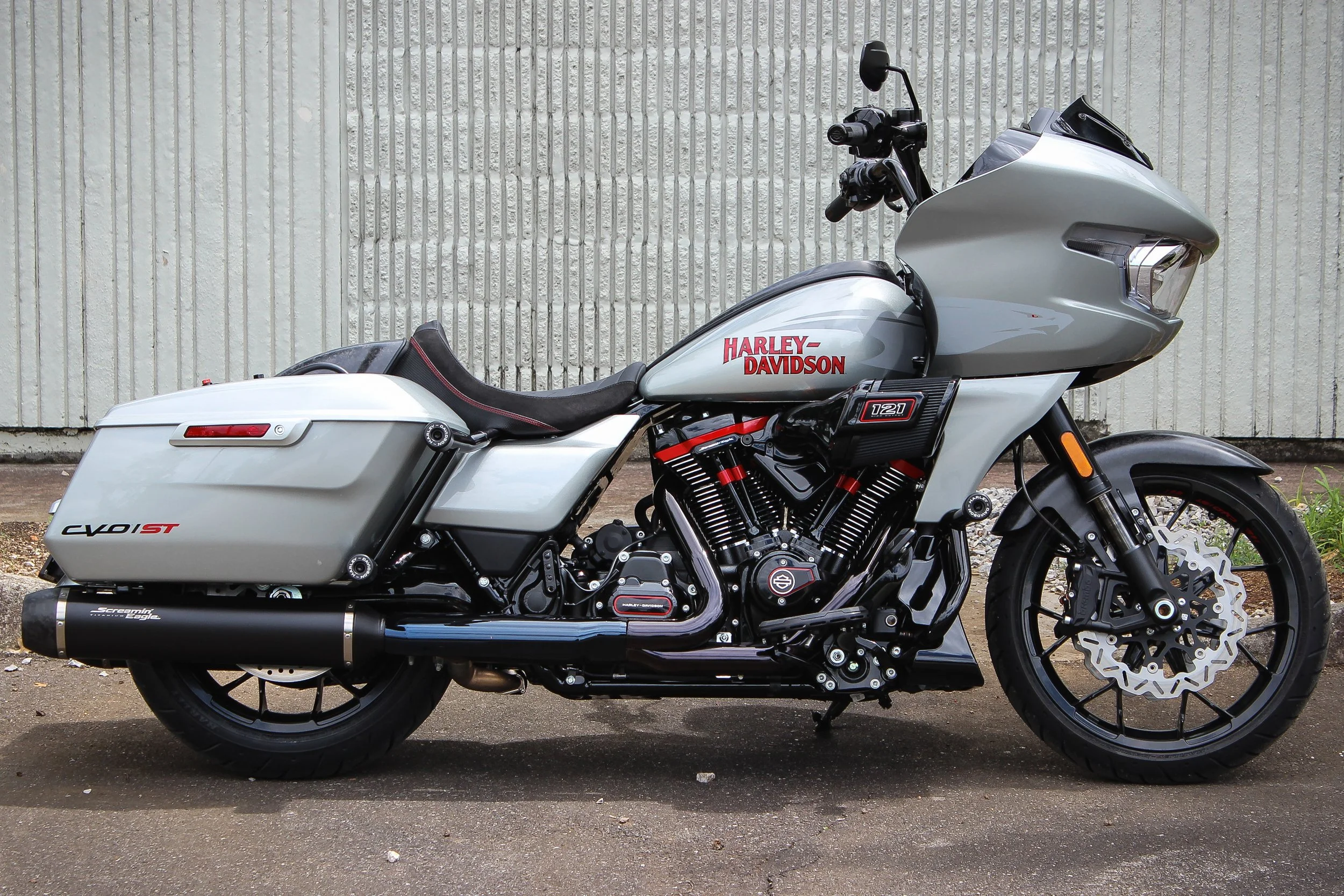 A Harley-Davidson touring motorcycle with a silver fairing, black and red accents, and hard saddlebags, parked on a street against a textured white wall background.