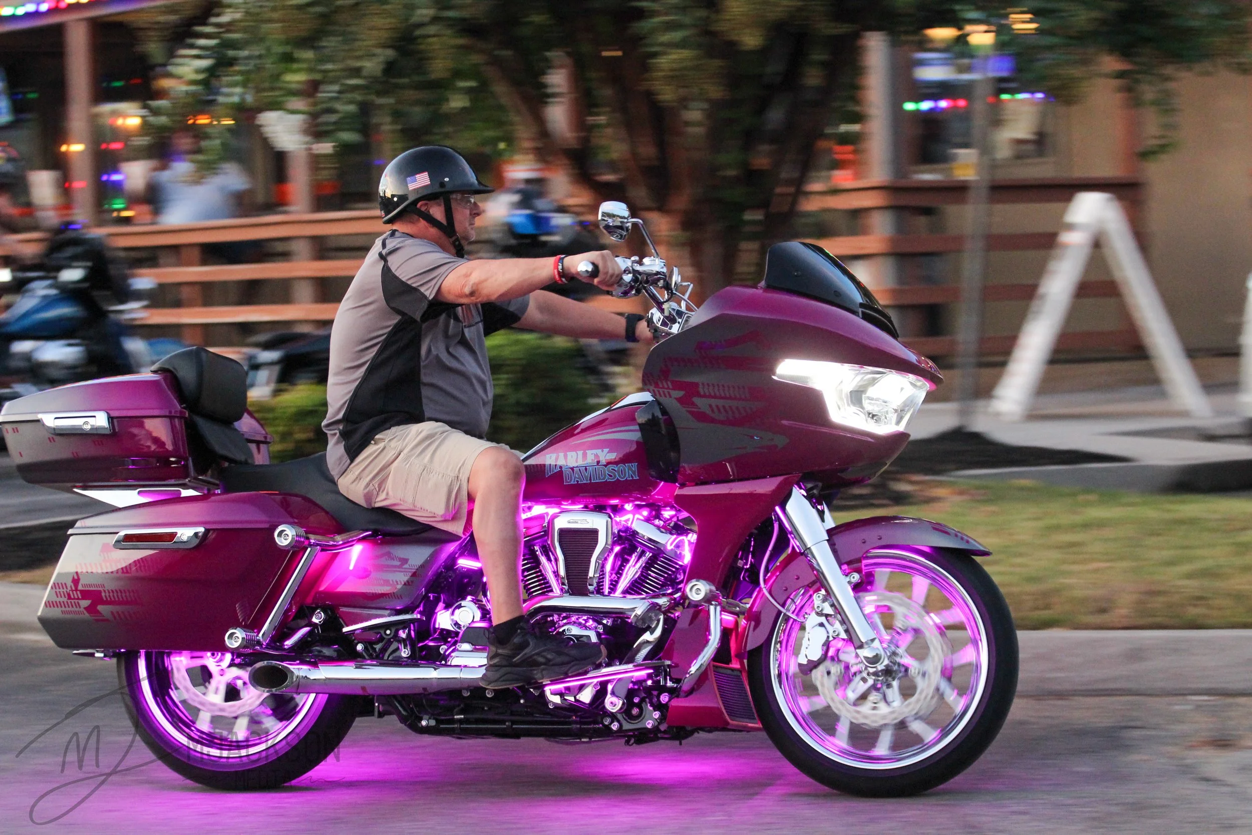 A man wearing a helmet and beige shorts riding a pink Harley Davidson motorcycle with purple neon lights underneath, driving through a street scene during evening or night.