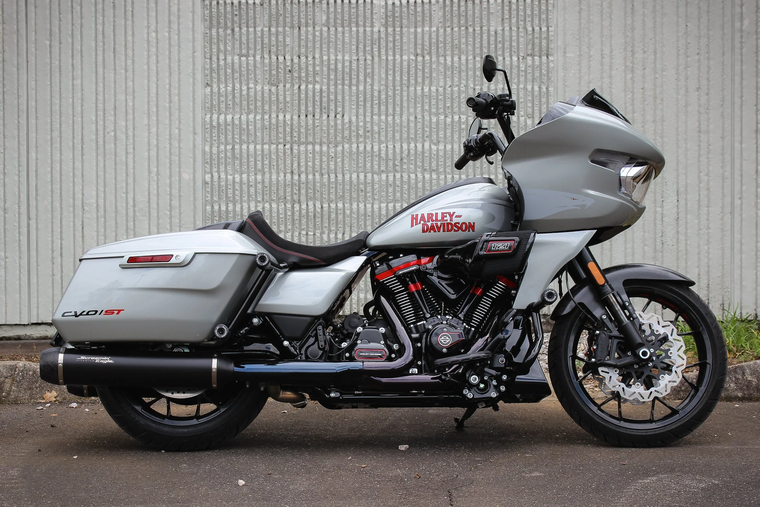 A silver Harley-Davidson motorcycle with saddlebags parked on the side of a street in front of a white textured wall.
