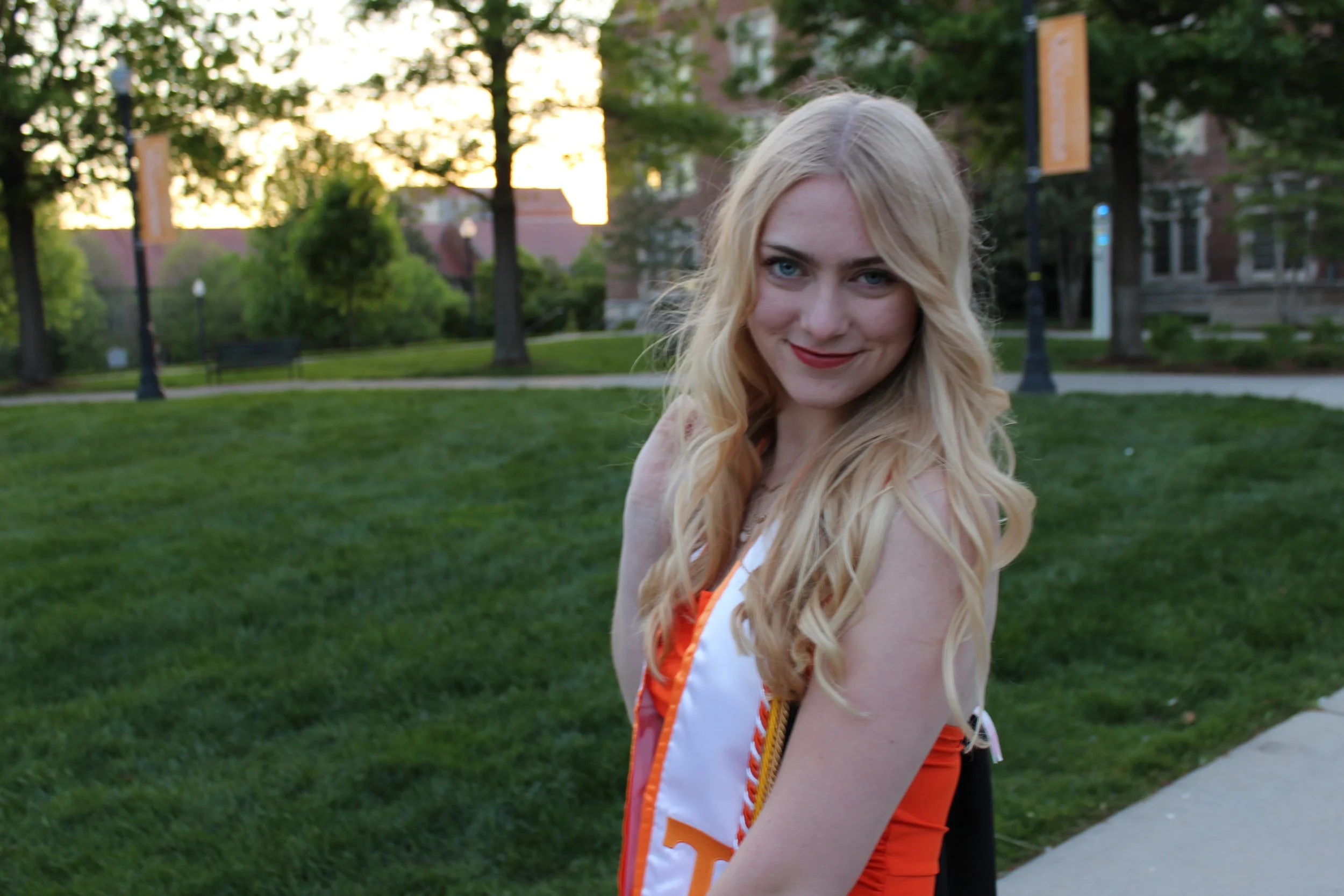 A young woman with long blonde hair and blue eyes smiling outdoors in a park during sunset, wearing a white and orange cheerleading uniform.
