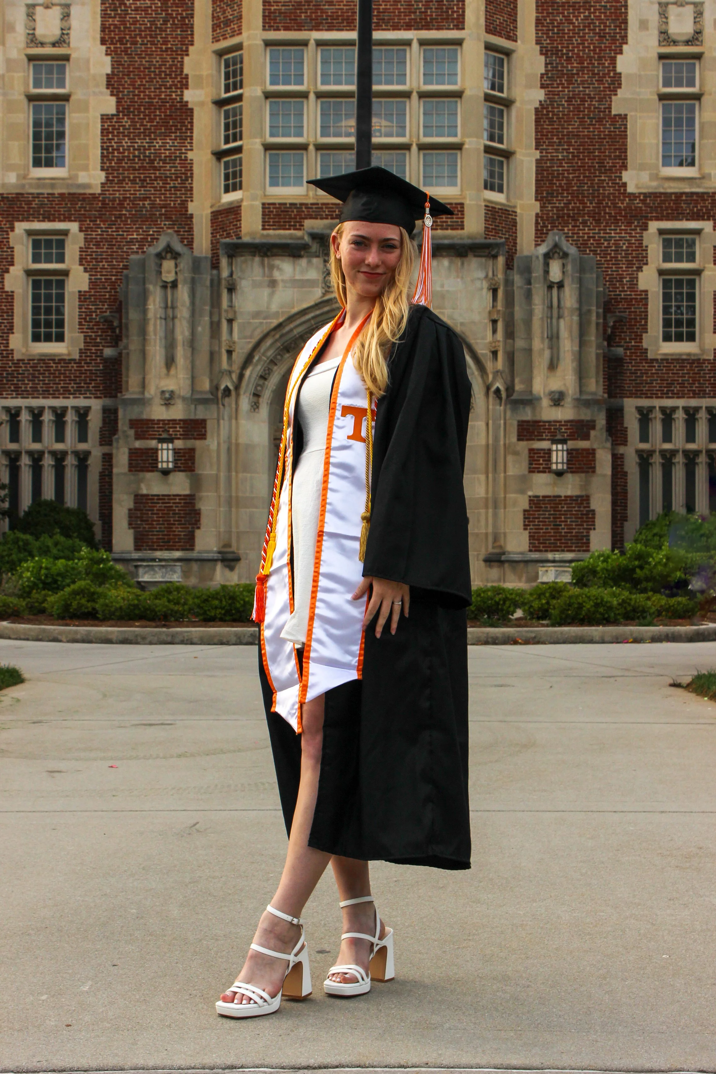 A young woman in a graduation cap and gown standing in front of a large brick building with decorative stonework. She is wearing graduation cords and white high-heeled sandals.