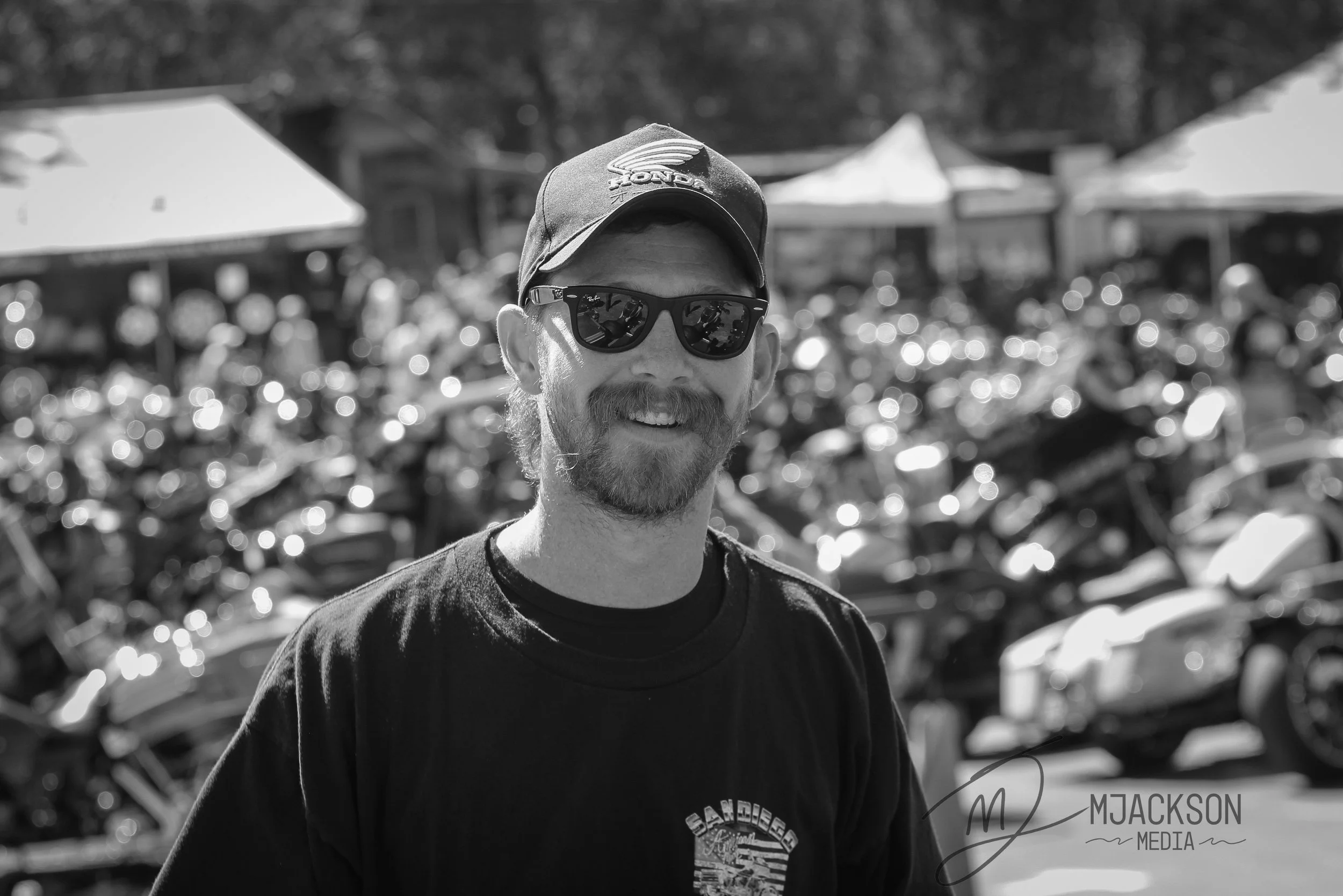 A man smiling wearing sunglasses and a baseball cap outdoors, with blurred tents or umbrellas in the background.
