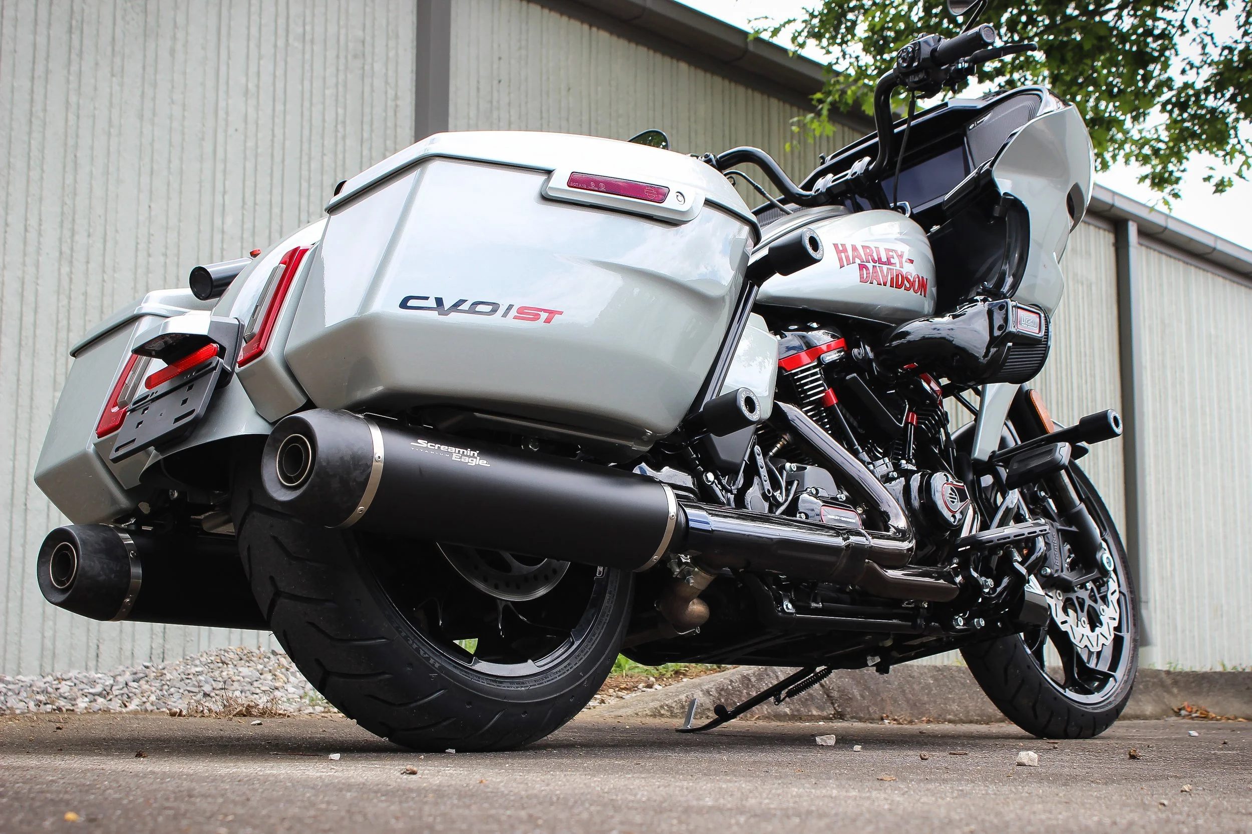 A white Harley-Davidson motorcycle parked on the ground with a large saddlebag, exhaust pipes, and a digital display, positioned outdoors near a gravel and asphalt surface.