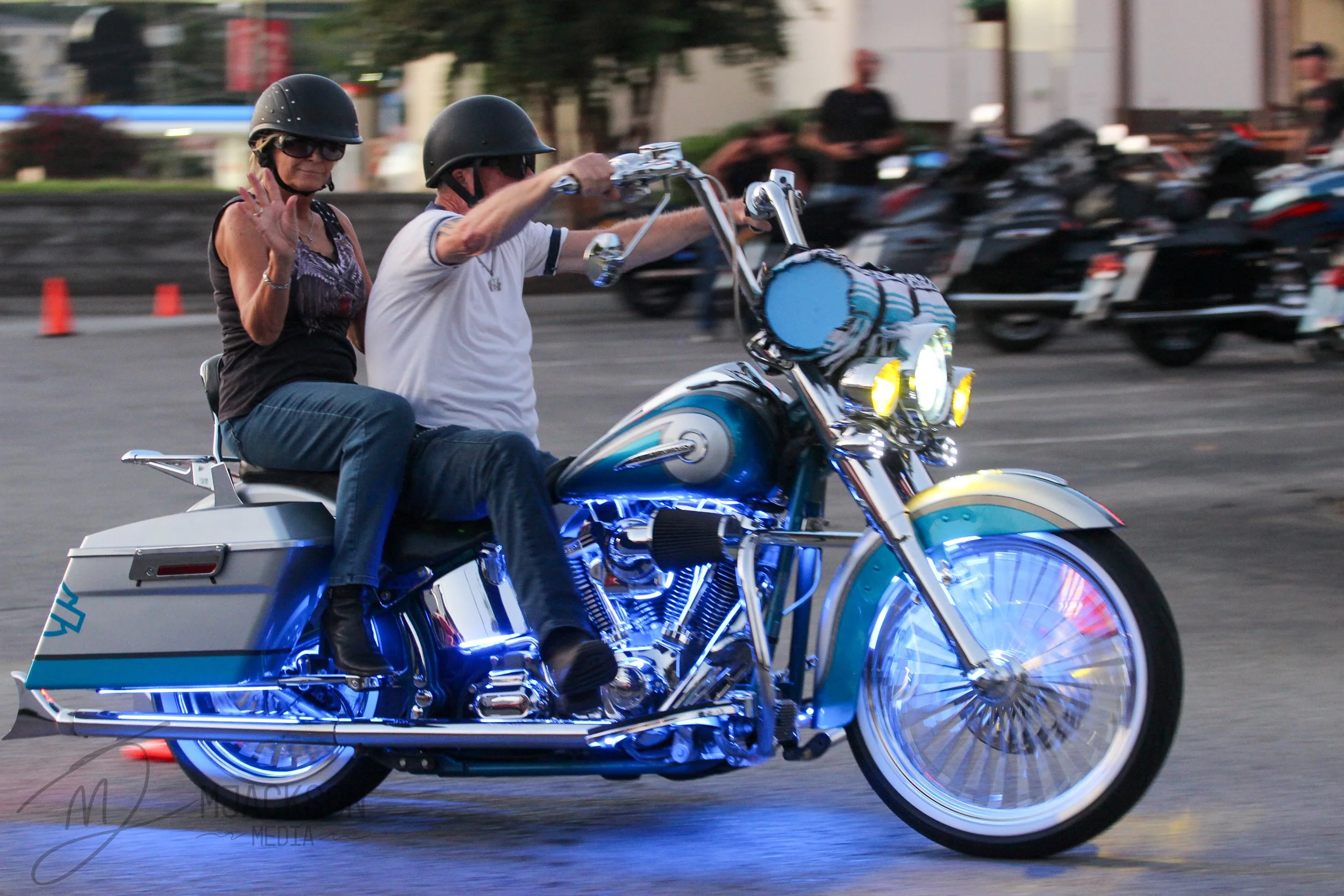 A man and woman riding a motorcycle at night, both wearing black helmets and casual clothes, with the woman waving and looking at the camera, in a parking lot with cars and cones in the background.