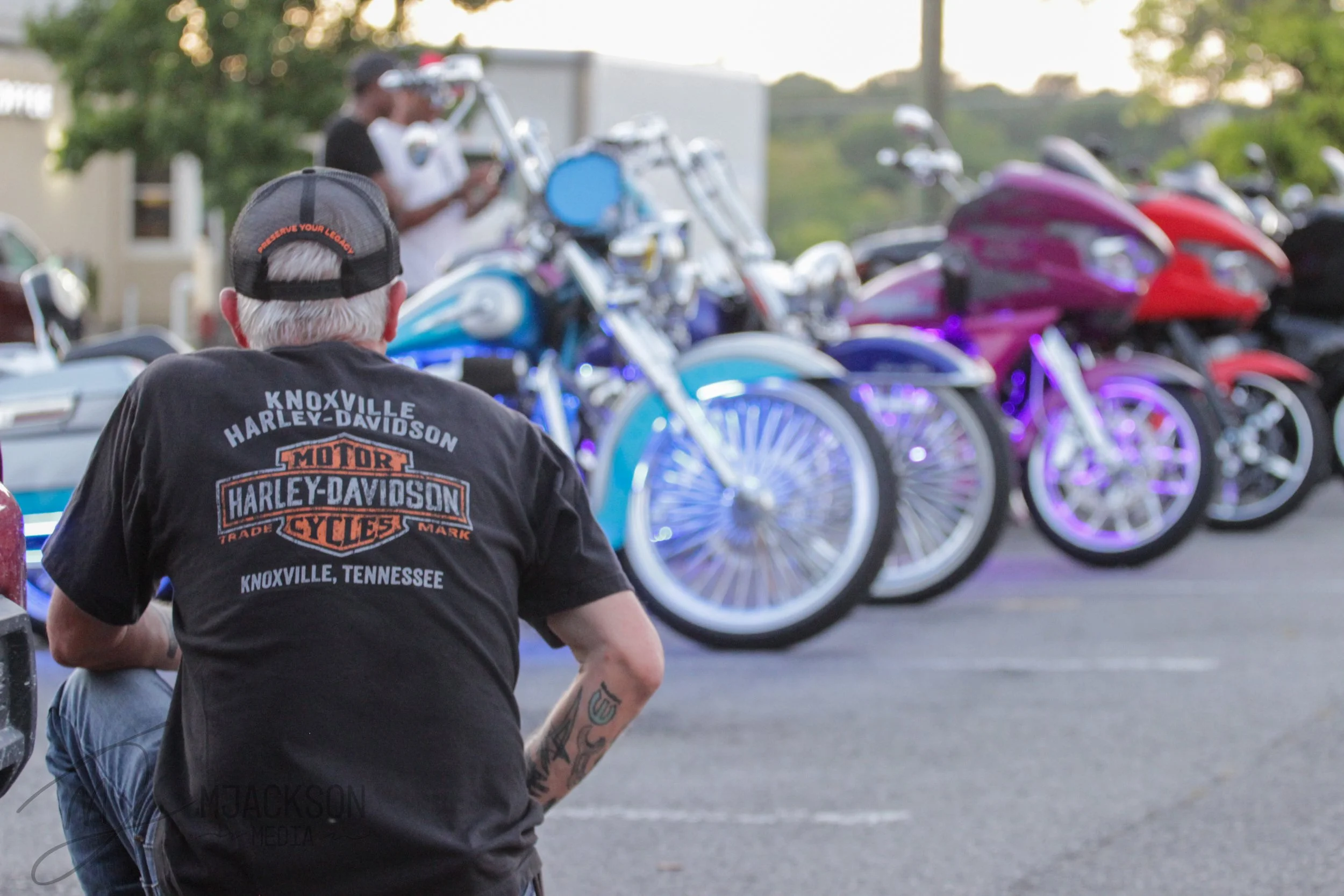 Person wearing a black Harley-Davidson Knoxville Tennessee T-shirt and a black cap jogging near a row of colorful motorcycles parked outdoors with green trees in the background.