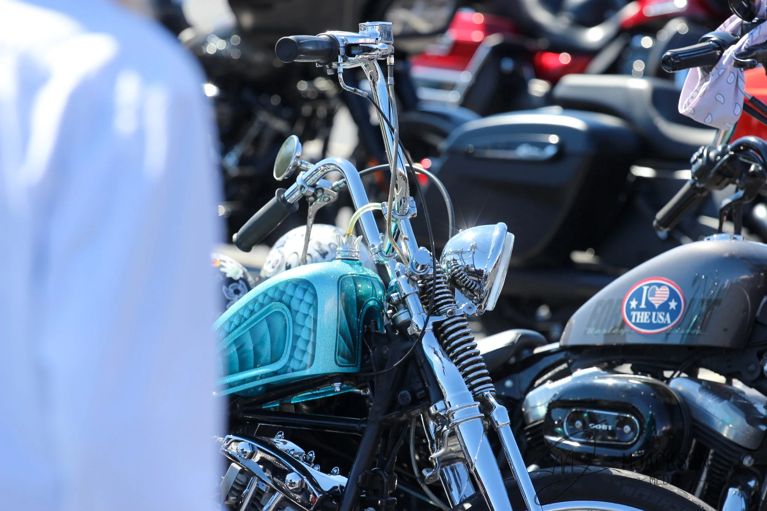 Close-up of a parked turquoise motorcycle with chrome details, surrounded by other motorcycles and scooters at a gathering or parking lot.