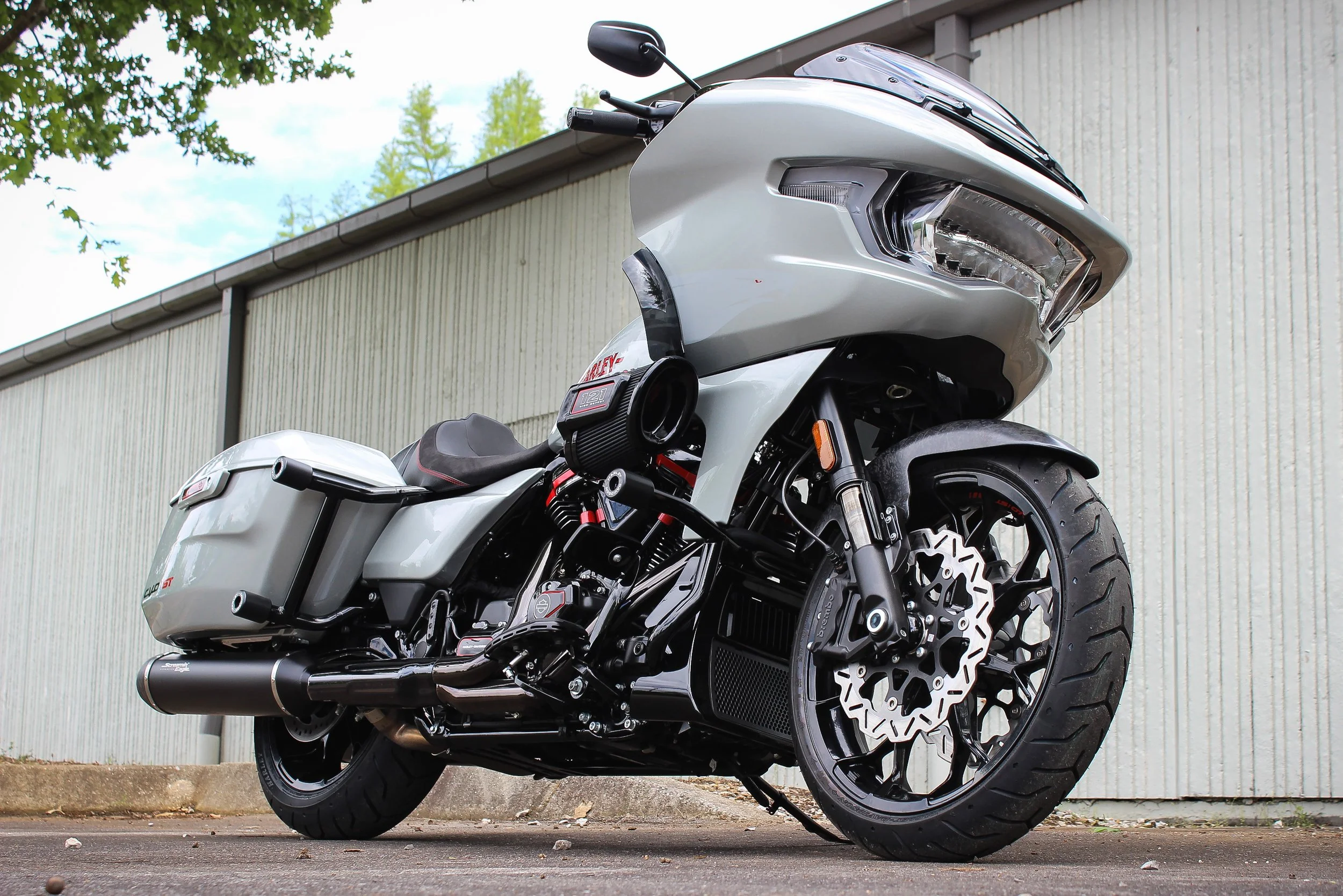 A white touring motorcycle parked outdoors on pavement, with a large front fairing, side storage compartments, and thick tires, against a background of a beige building and green trees.