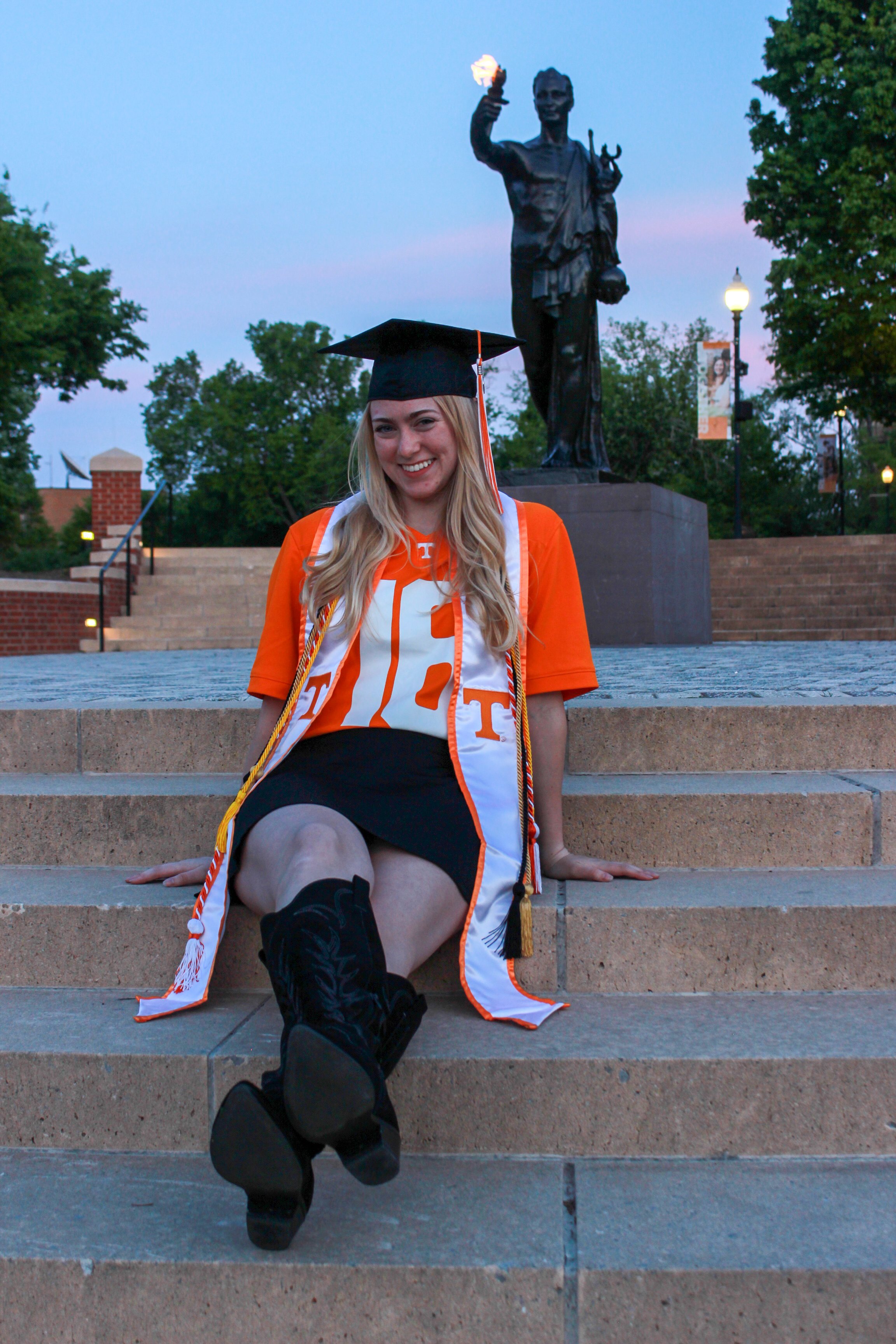 A young woman in a graduation cap and gown, smiling, sitting on steps outside. Behind her is a statue holding a torch, surrounded by trees and lampposts, during dusk.