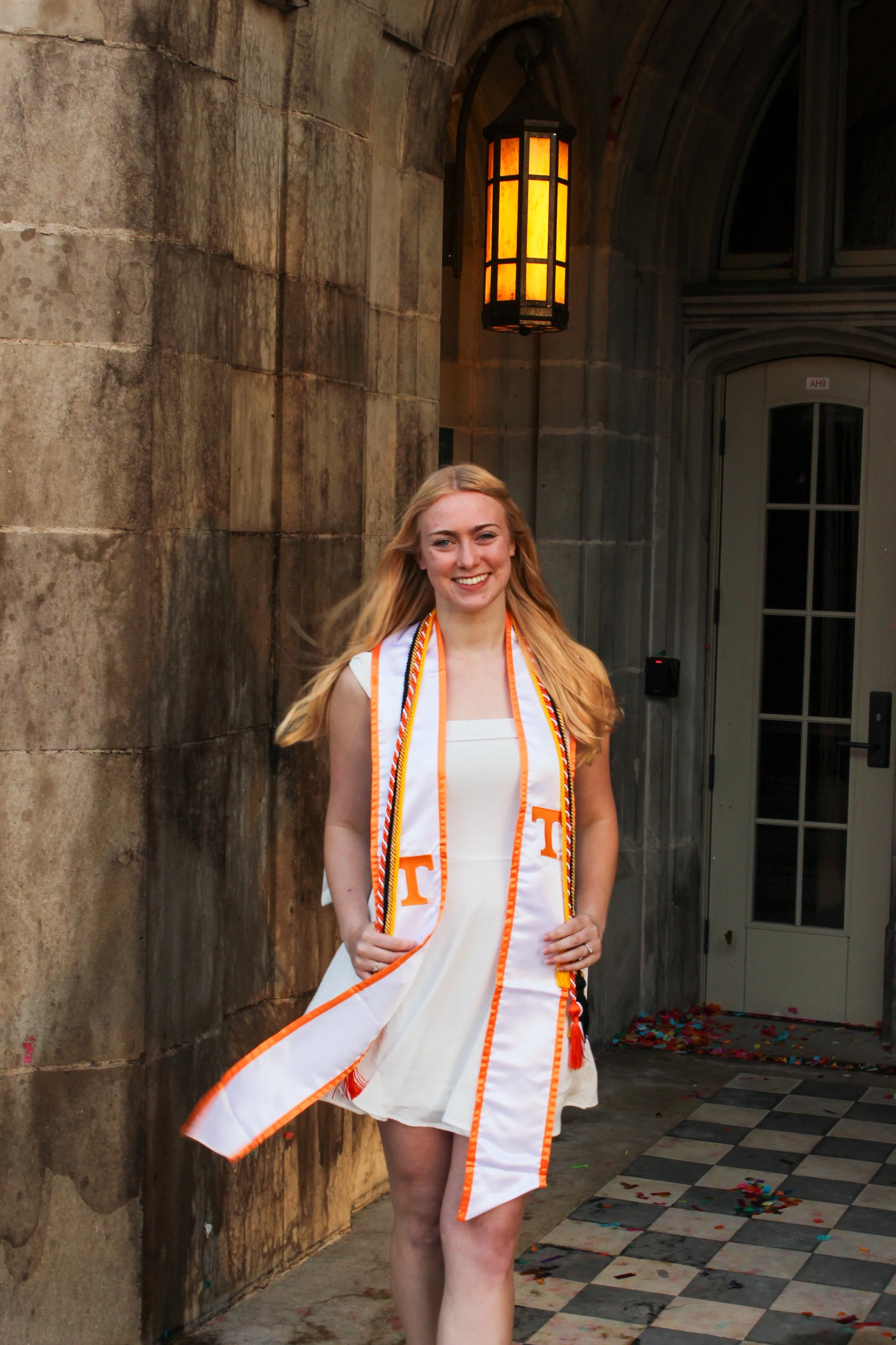 Young woman with long blonde hair wearing a white dress and graduation stole, smiling, standing outside near stone walls and a decorative lantern, with confetti on the ground.