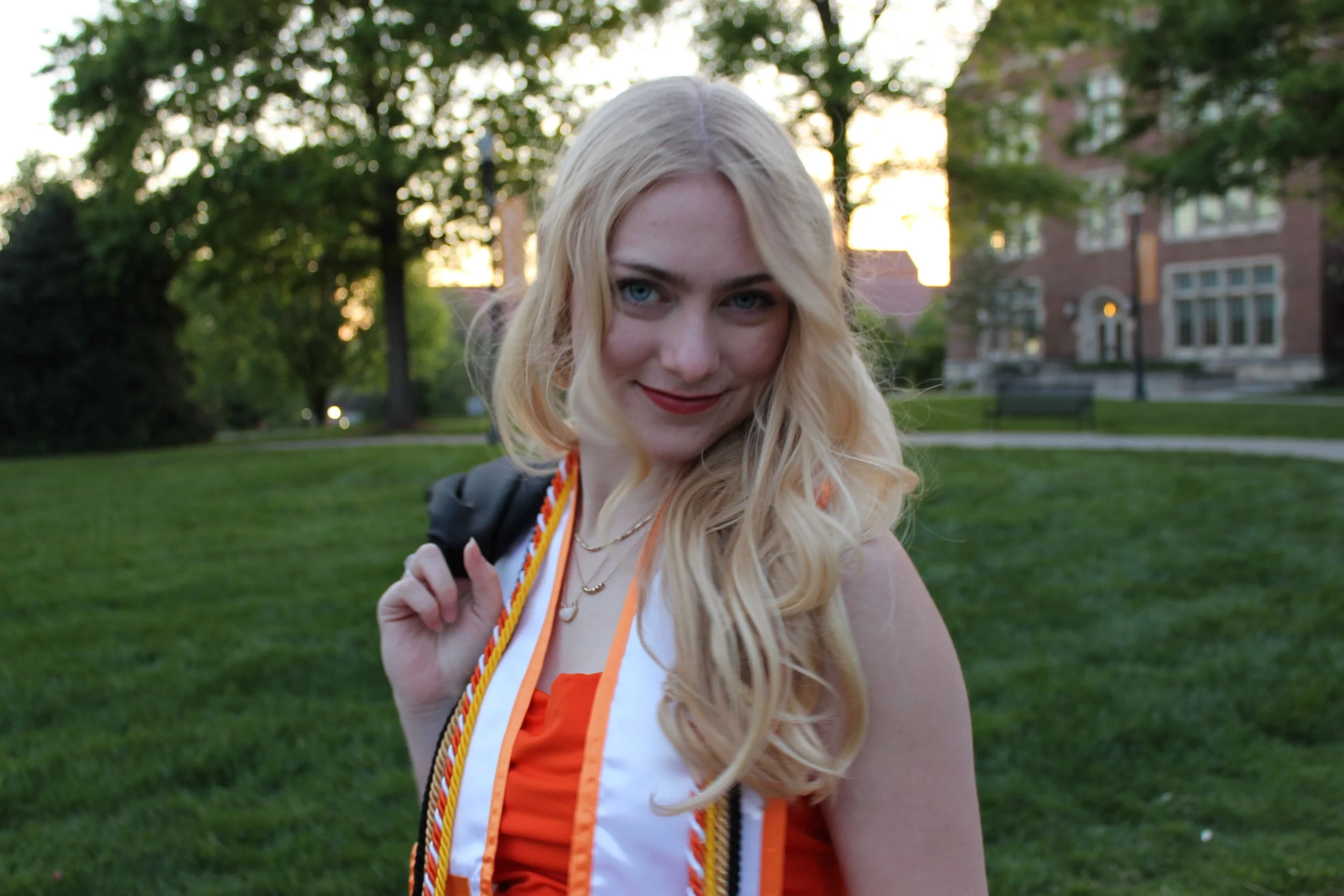 Young woman with blonde hair smiling outdoors holding a graduation cord, background includes trees and a brick building at sunset.