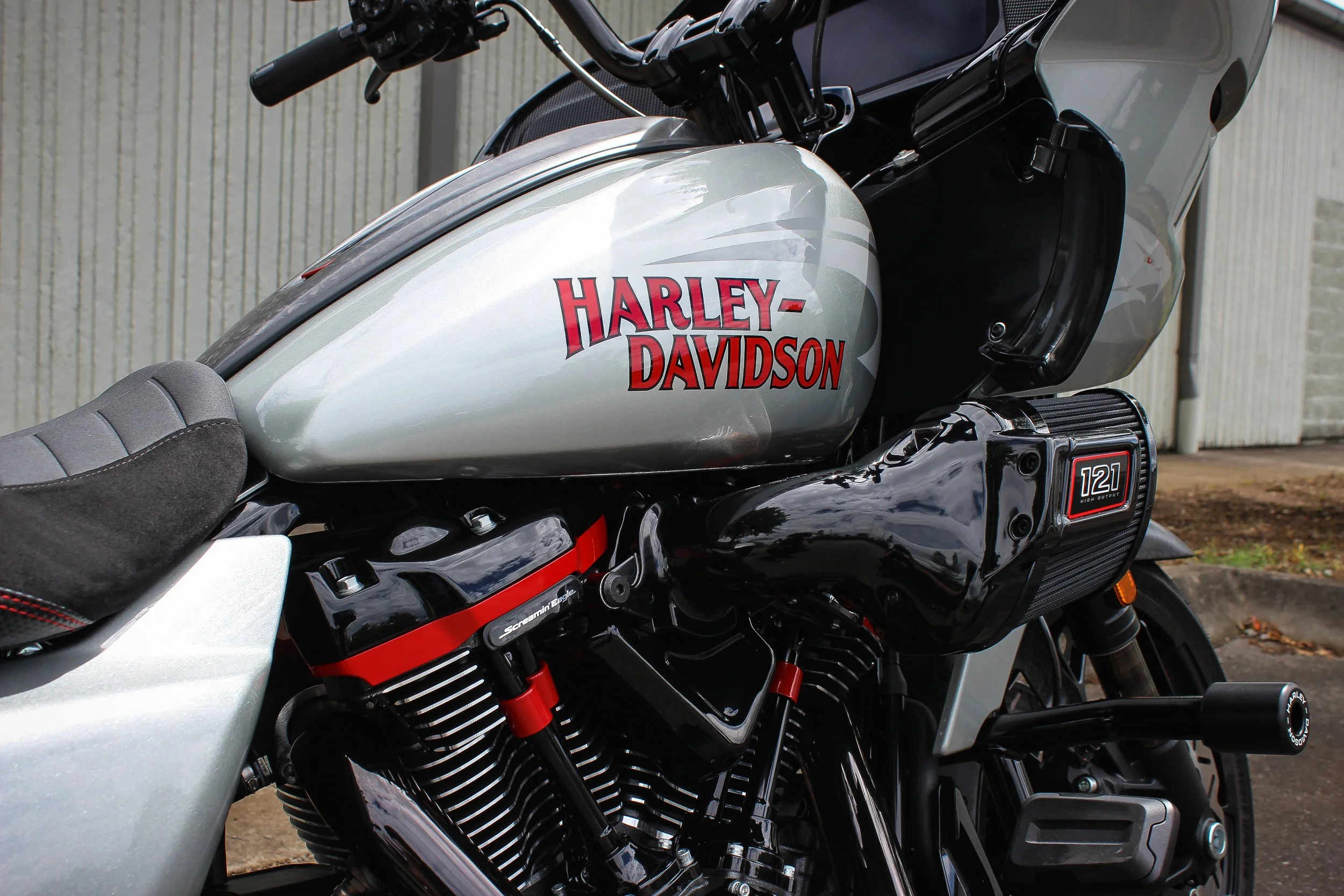 Close-up of a Harley-Davidson motorcycle's fuel tank with the brand logo, partially showing the handlebars, engine, and seat, parked against a backdrop of a fence and trees.