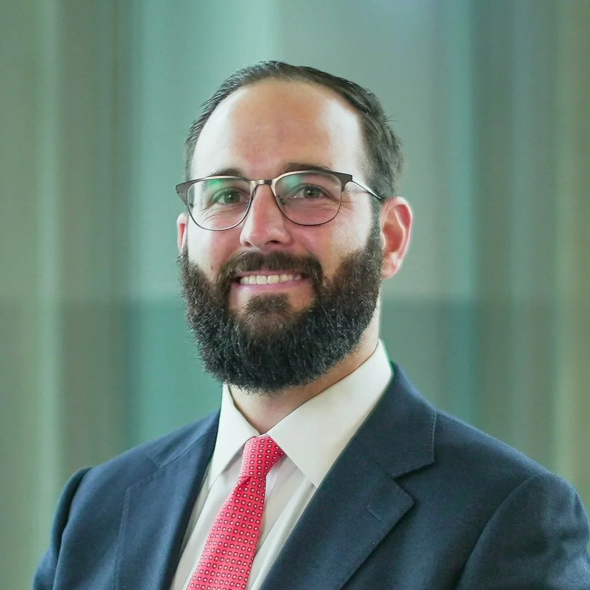 A professional man with glasses, a full beard, in a navy suit, white shirt, and pink tie, smiling, with a blurred office background.