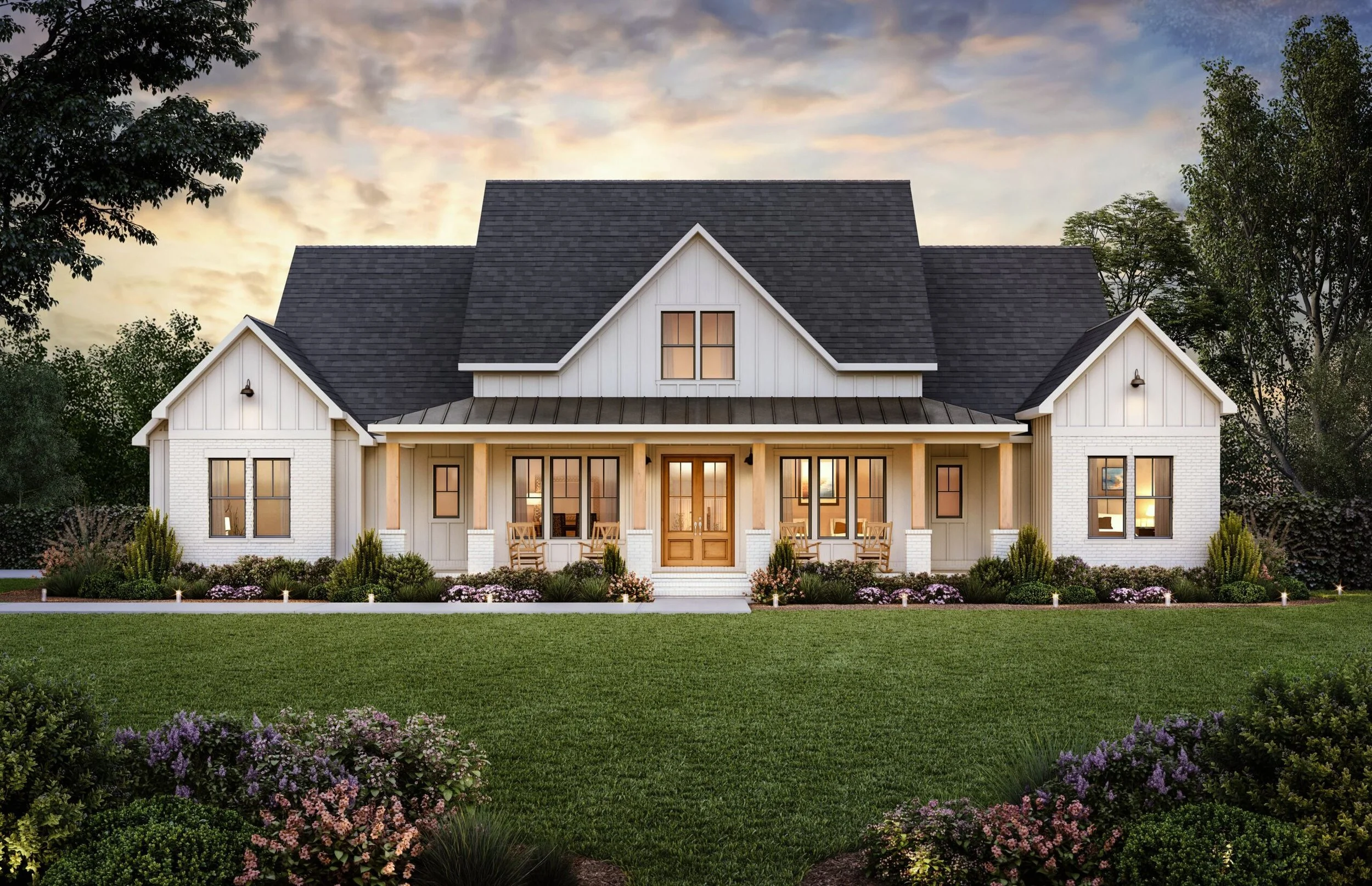 A large, modern house with white exterior walls, dark gray roof, front porch with wooden columns, and landscaped front yard with grass, bushes, and flowers during sunset.