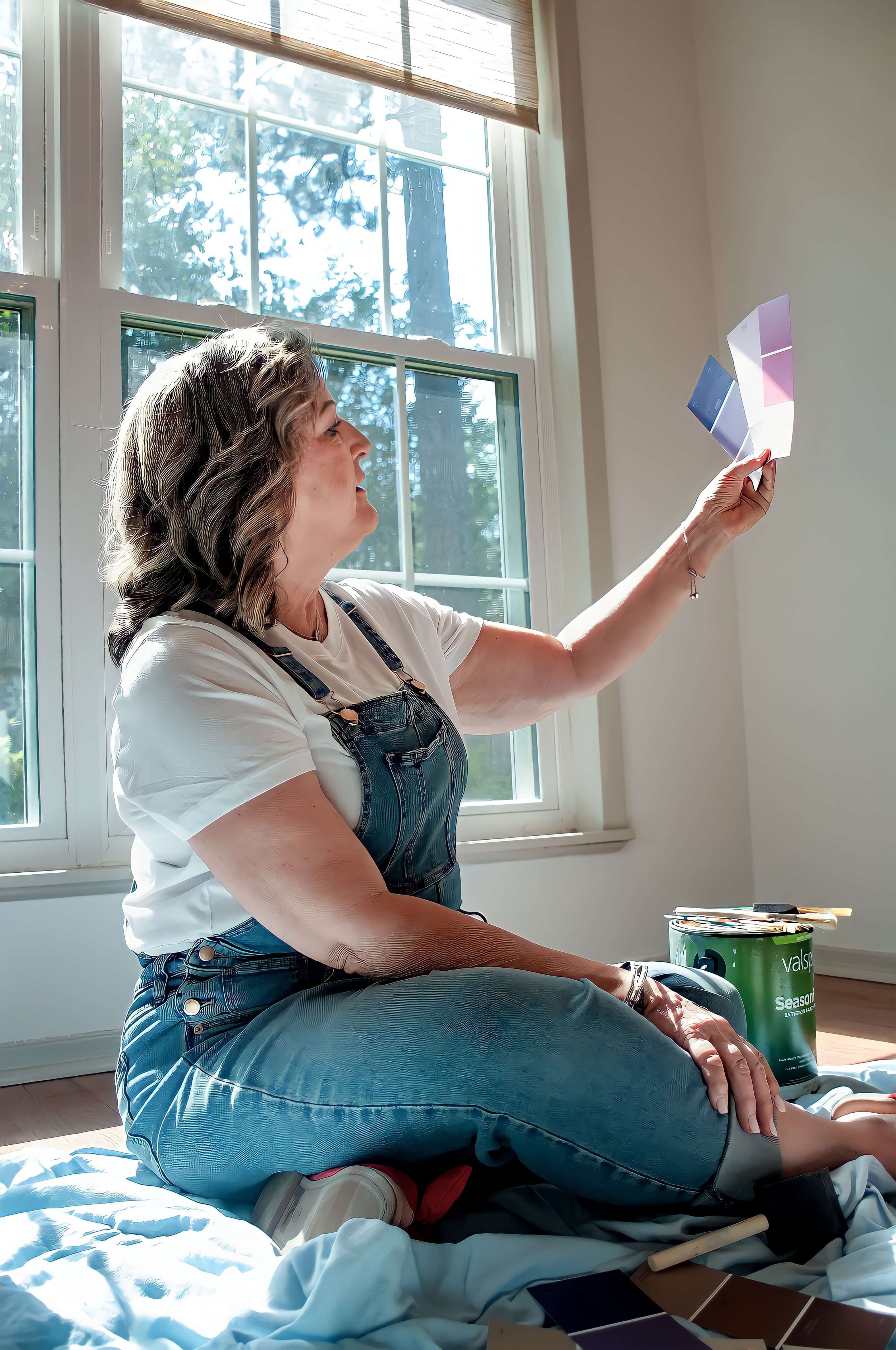 Sherri Davis sitting on a white sheet on the floor, holding up color swatches or paint samples near a window with sunlight streaming in.