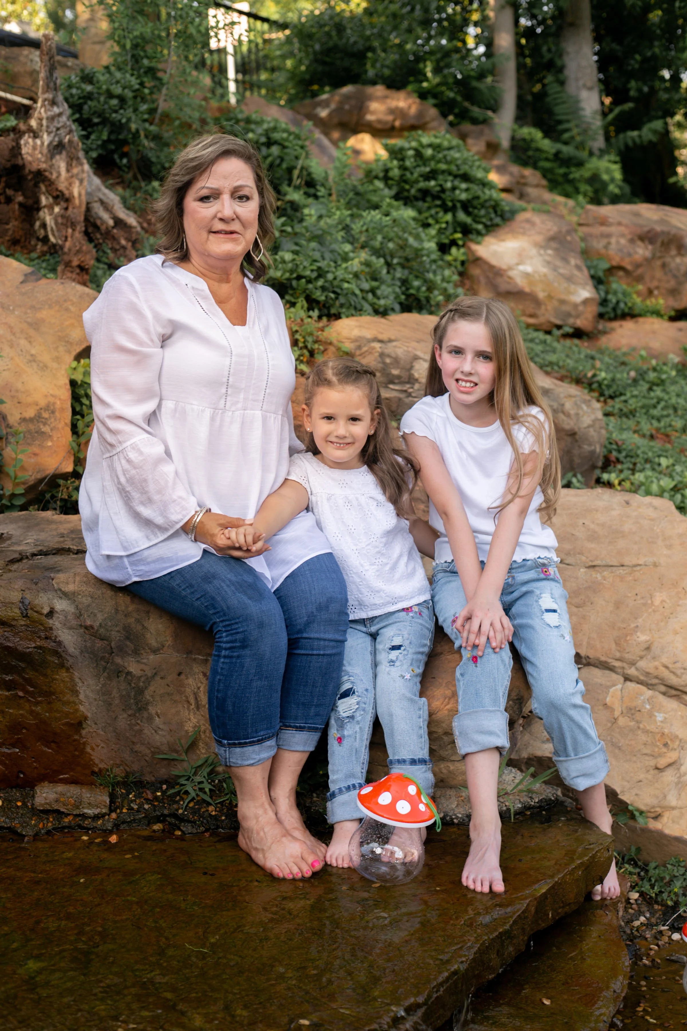 Sherri Davis with her two young granddaughters sitting outdoors on rocks near a small stream in a lush garden.