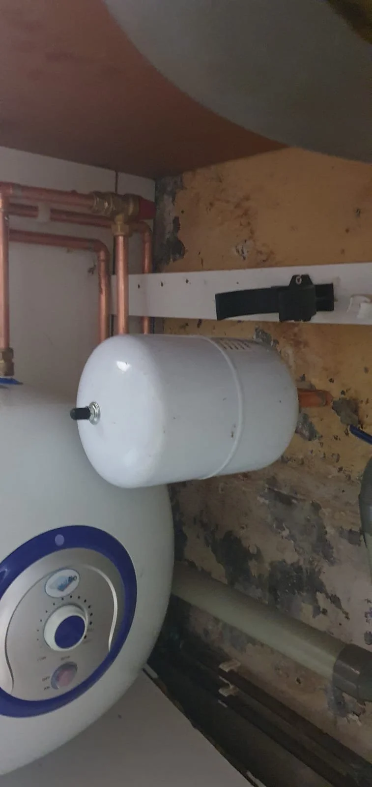 White electric water heater with attached expansion tank, copper plumbing, and wall-mounted bracket in a utility room