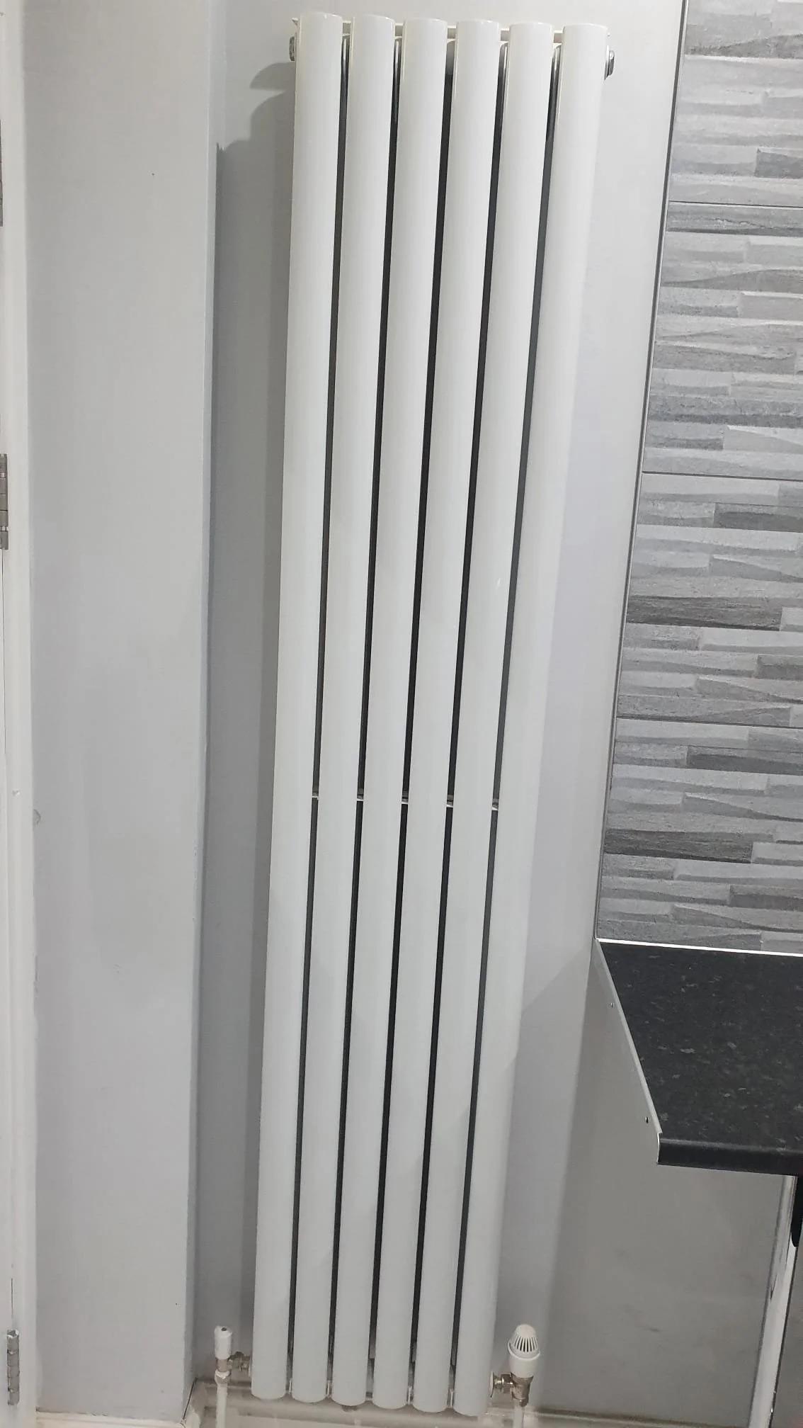 Tall white vertical panel radiator mounted on a gray tiled wall beside a black countertop
