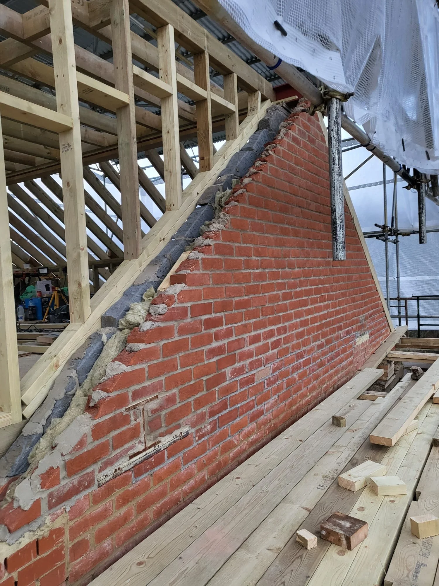 Brick wall and exposed timber roof framing under construction, surrounded by scaffolding and temporary weather protection.