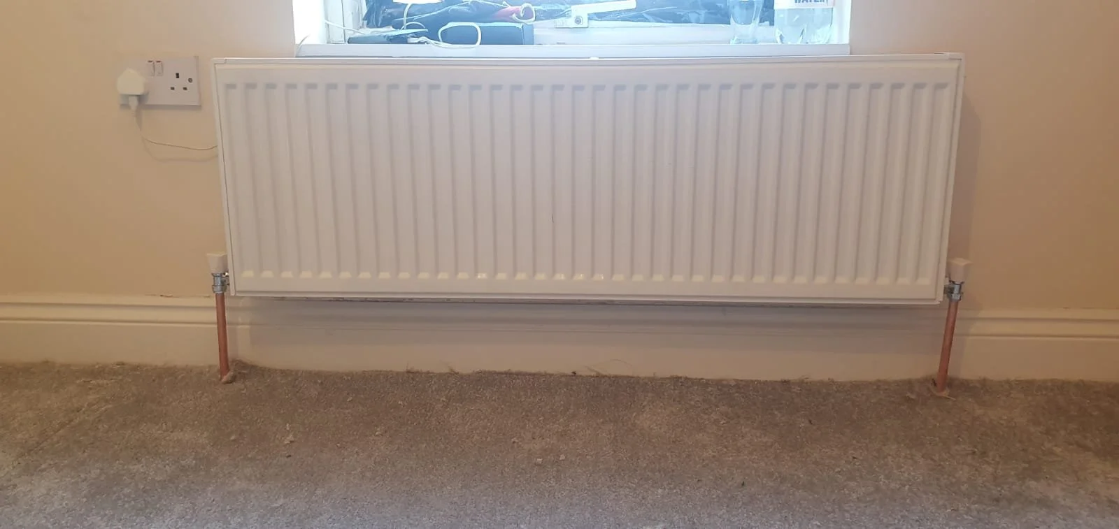 White wall-mounted radiator beneath a window, with exposed copper pipes and valves above a carpeted floor.