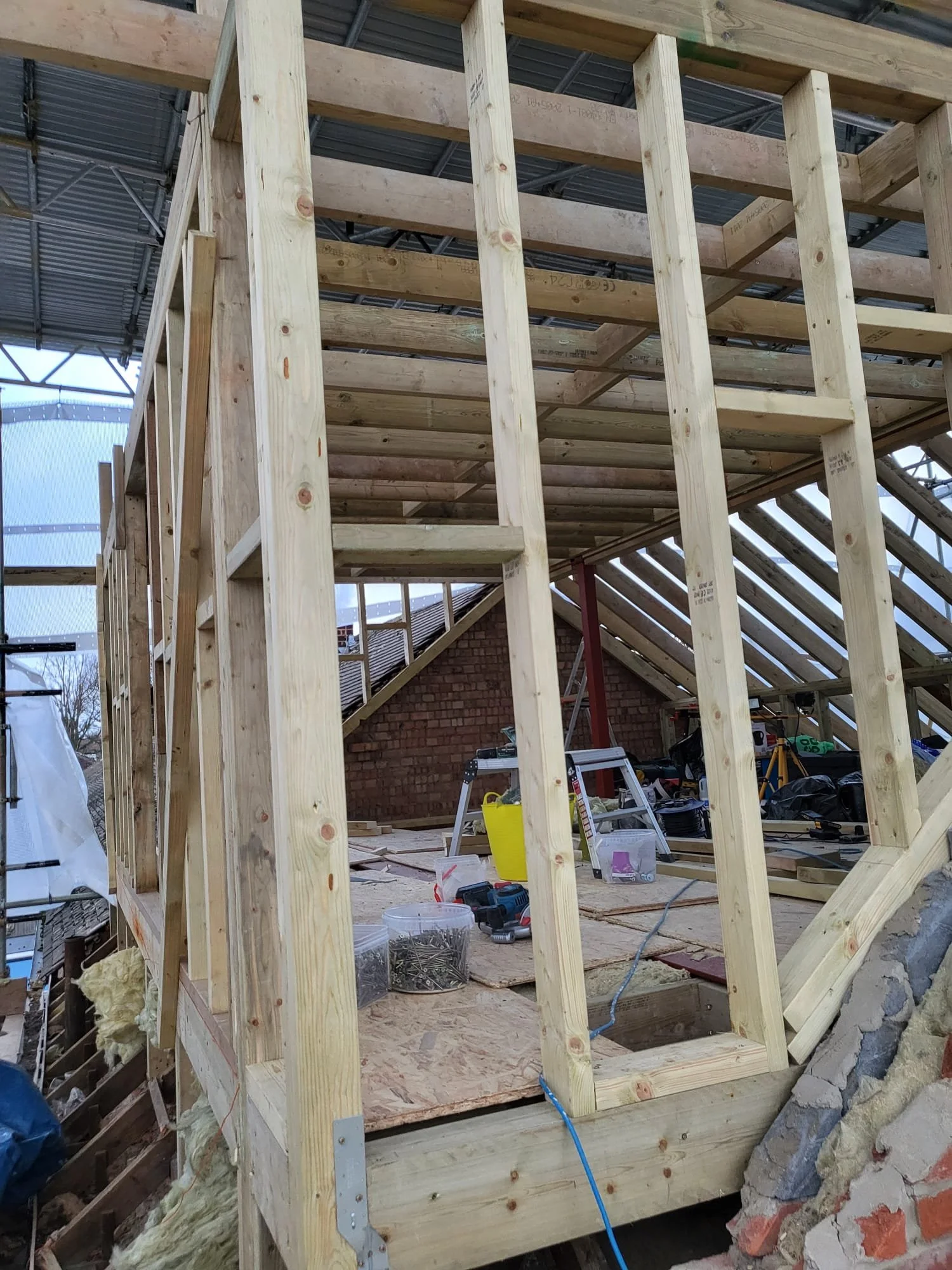 Wood-framed building under construction with exposed studs, roof beams, tools, ladders, insulation, and construction materials.