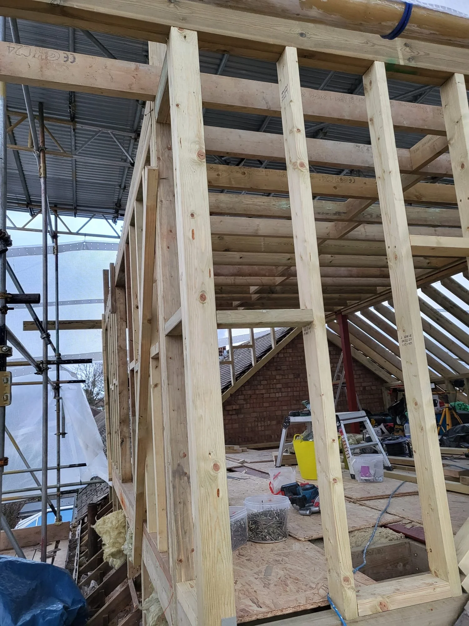 Timber-framed house extension under construction with exposed studs, roof beams, scaffolding, tools, and building materials