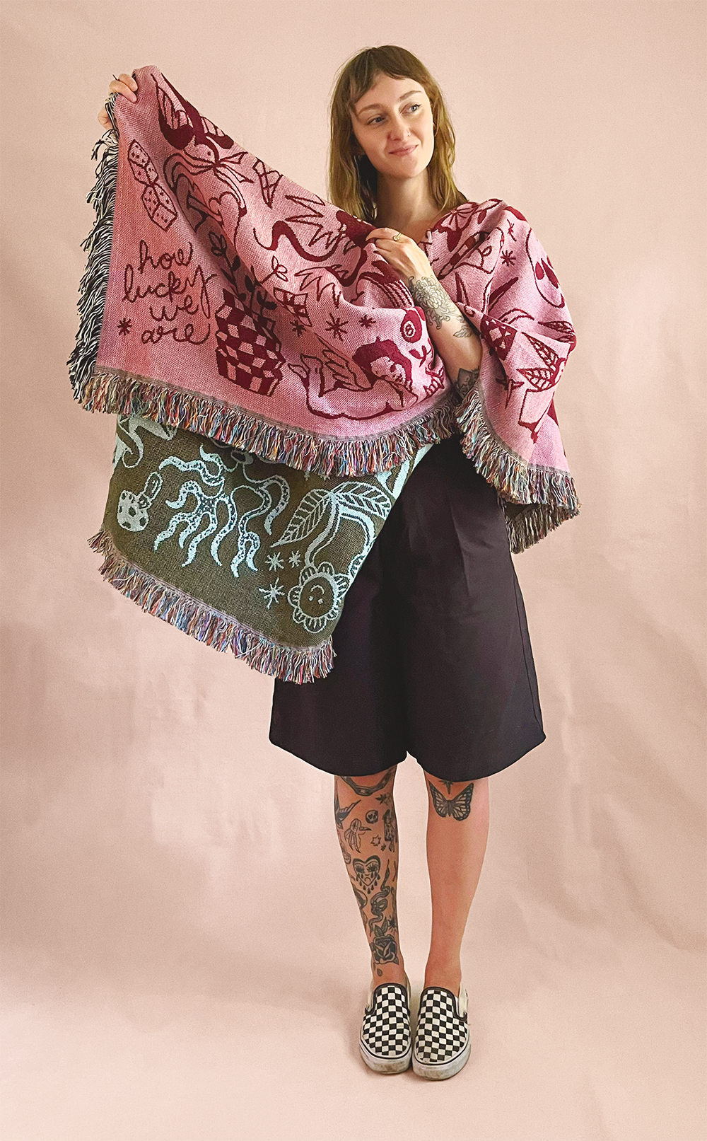 A woman with tattoos on her legs and arms, wearing a black skirt and colorful checkered slip-on shoes, holds two decorative blankets with abstract and animal designs, against a pink background.