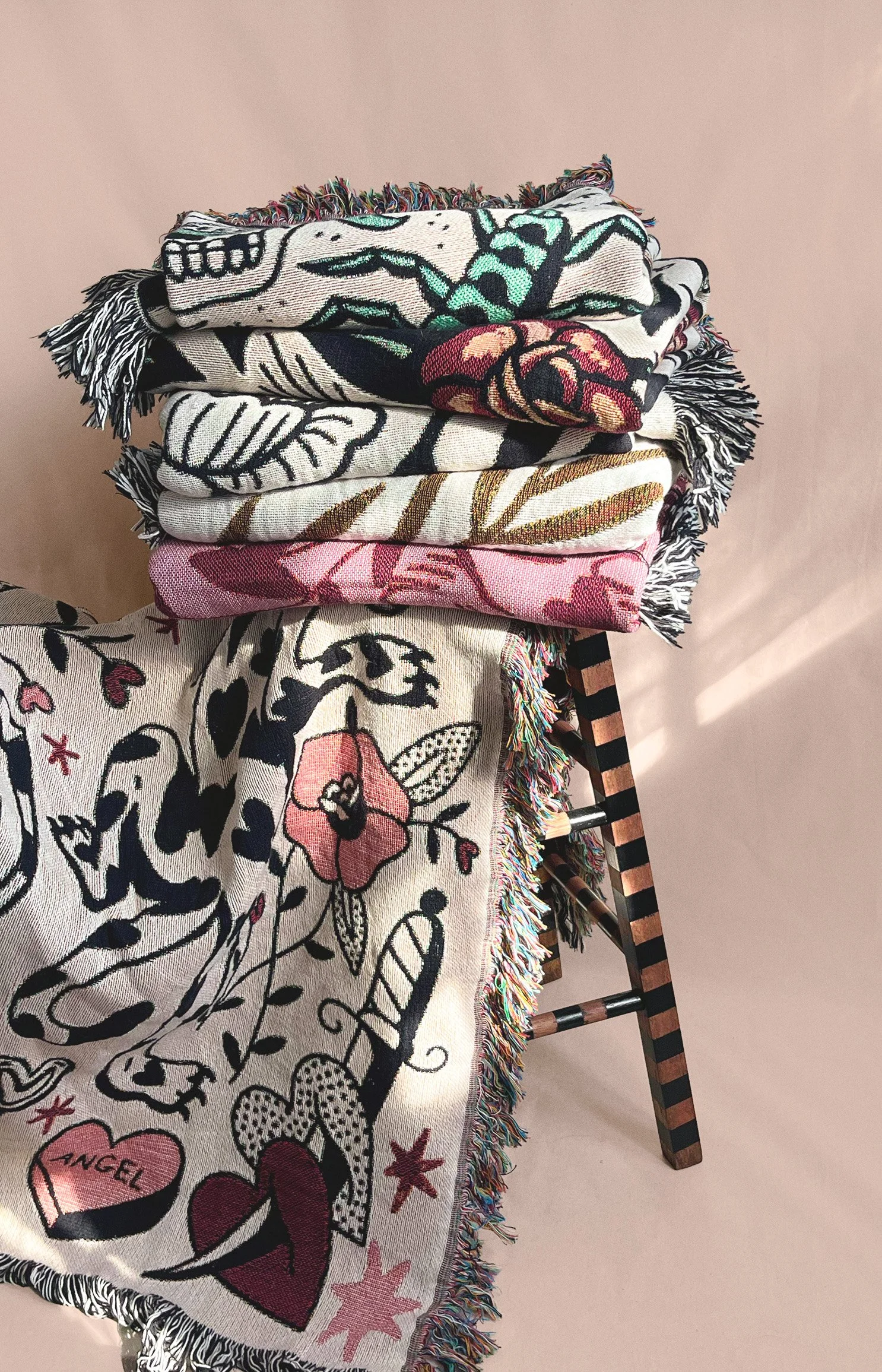 Stack of woven cotton blankets fringed edges placed on a wooden stool with black and brown stripes, against a plain light beige background.