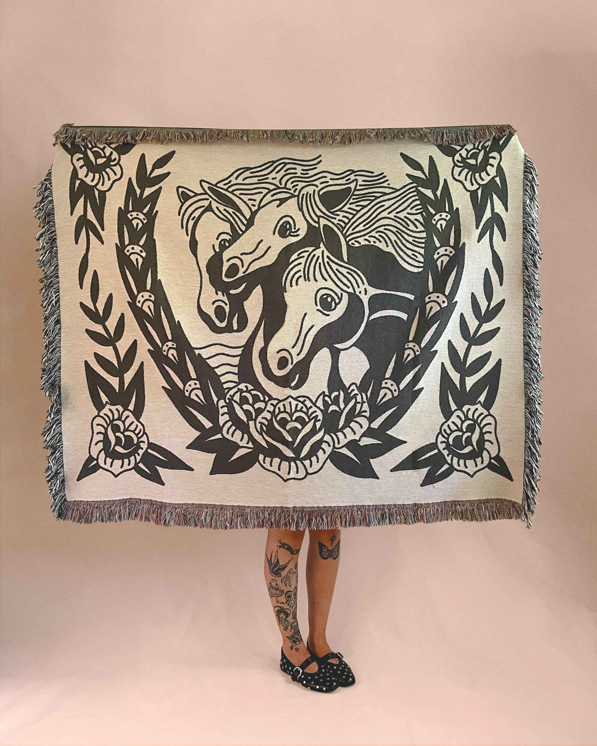 A person holding up a tapestry with a black and white illustration of two horses surrounded by floral and leaf patterns, with their legs and feet visible below the tapestry.