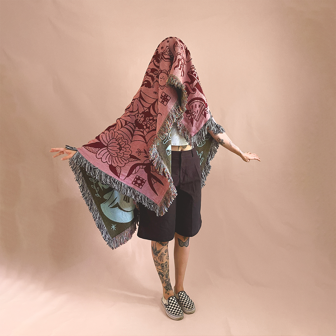 Person standing with their head covered by a large, decorative, colorful shawl with floral and other patterns, against a plain pink background, wearing black shorts, checkered slip-on shoes, and visible tattoos on their legs and arms.
