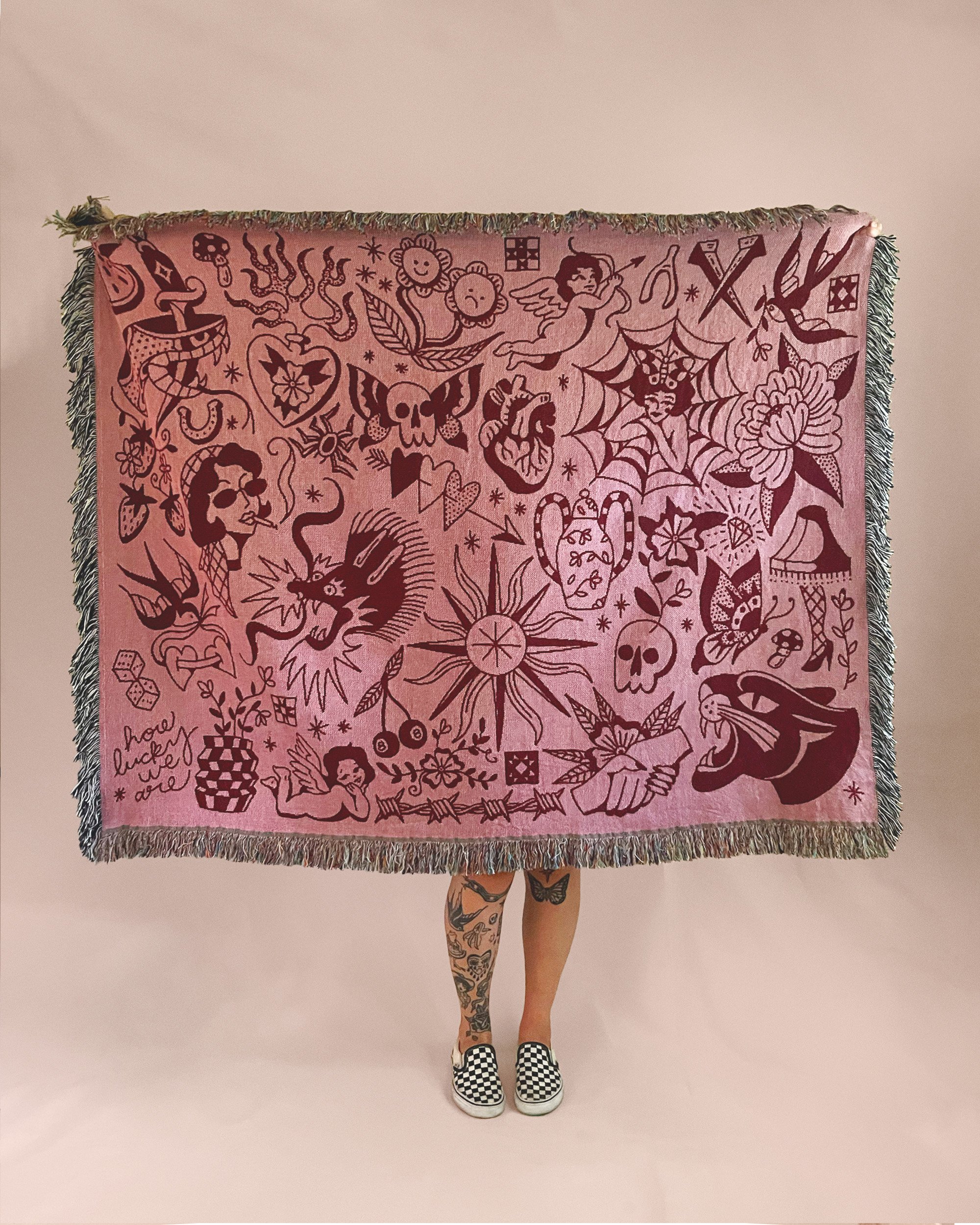 Person holding up a pink tattooed cloth with various black tattoo-style illustrations, wearing checkered slip-on shoes, and standing against a plain pink background.