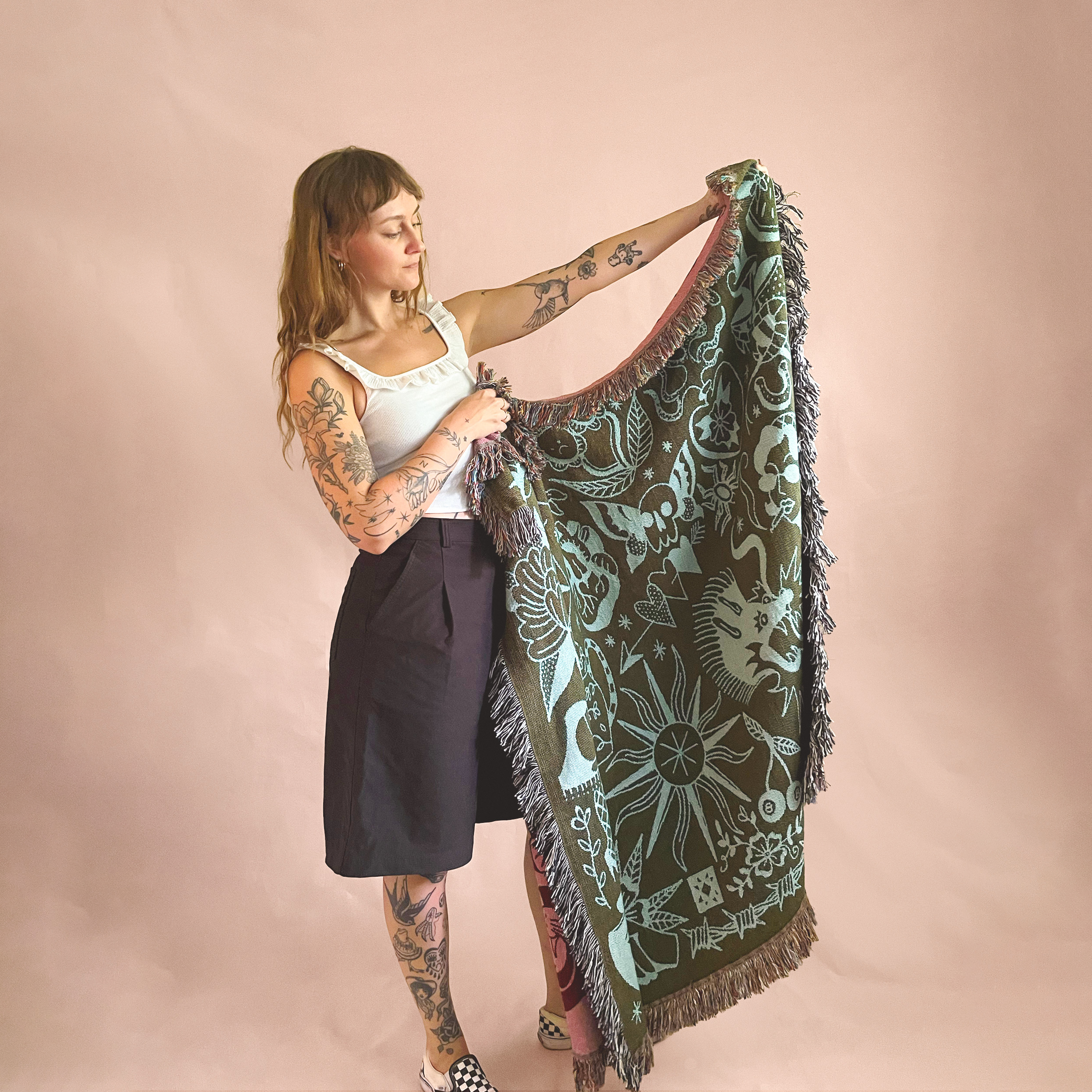 A woman with tattoos on her arms and legs, wearing a white sleeveless top, holding a large patterned textile against a pink background.