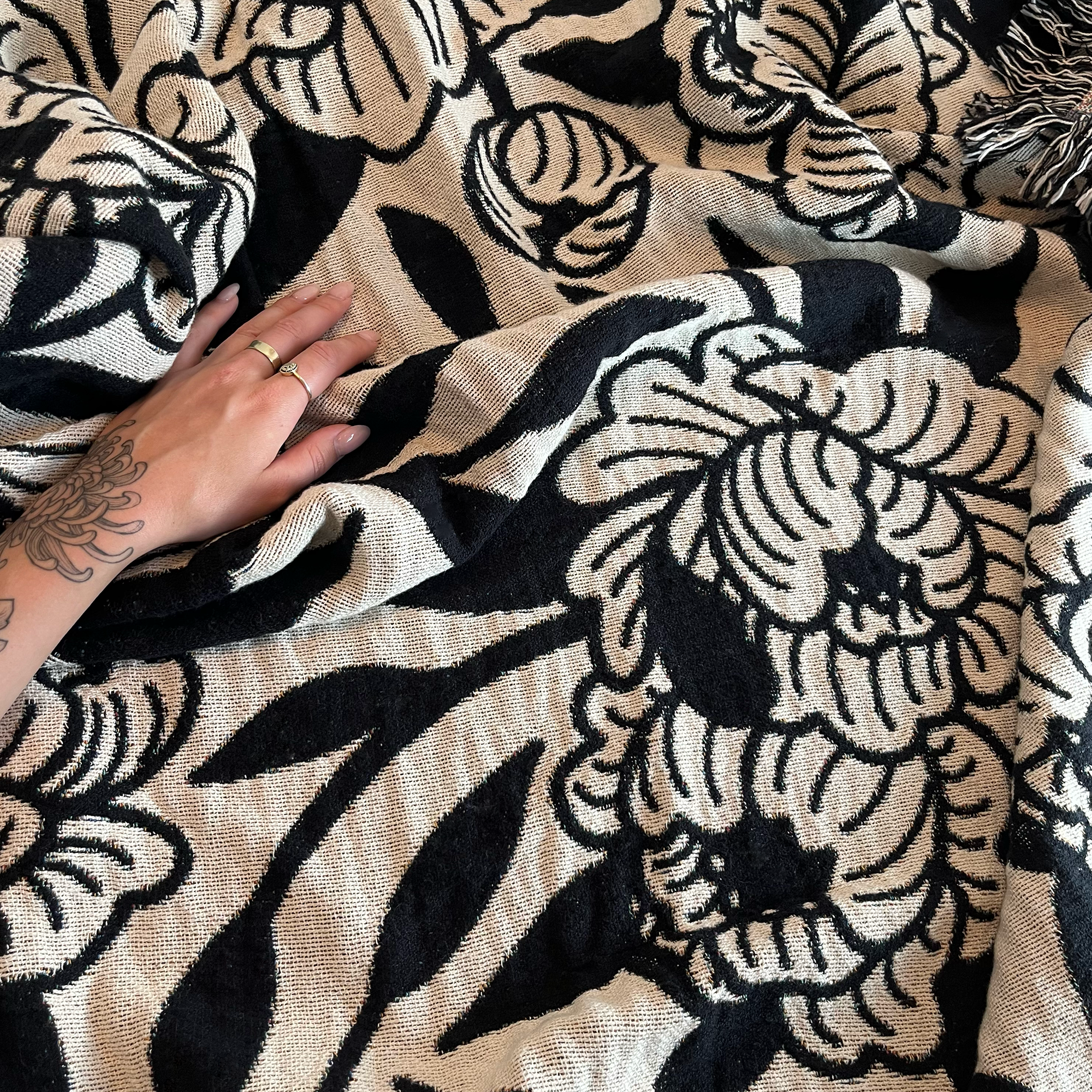 A person with a tattooed hand is resting on a black and beige patterned textile with floral and abstract designs, and fringed edges.