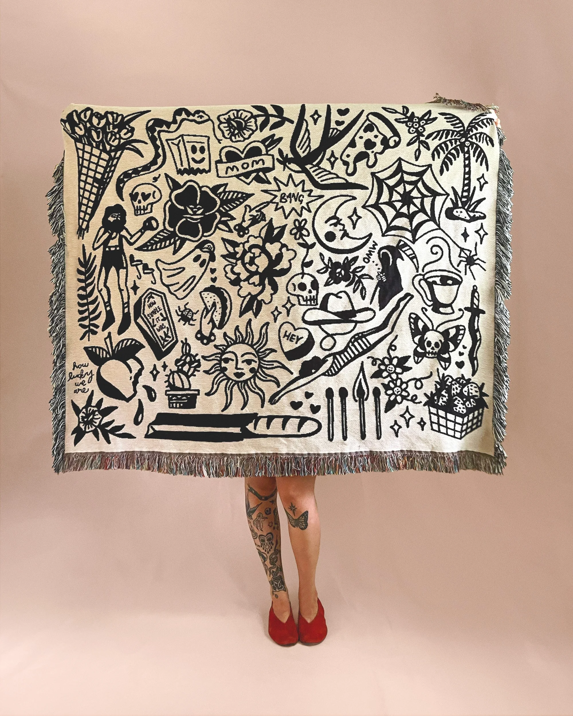 Person standing with a large blanket featuring black and white doodles and illustrations, including flowers, skulls, a sun, a moon, animals, and various symbols, with the person's legs and red shoes visible beneath the blanket.