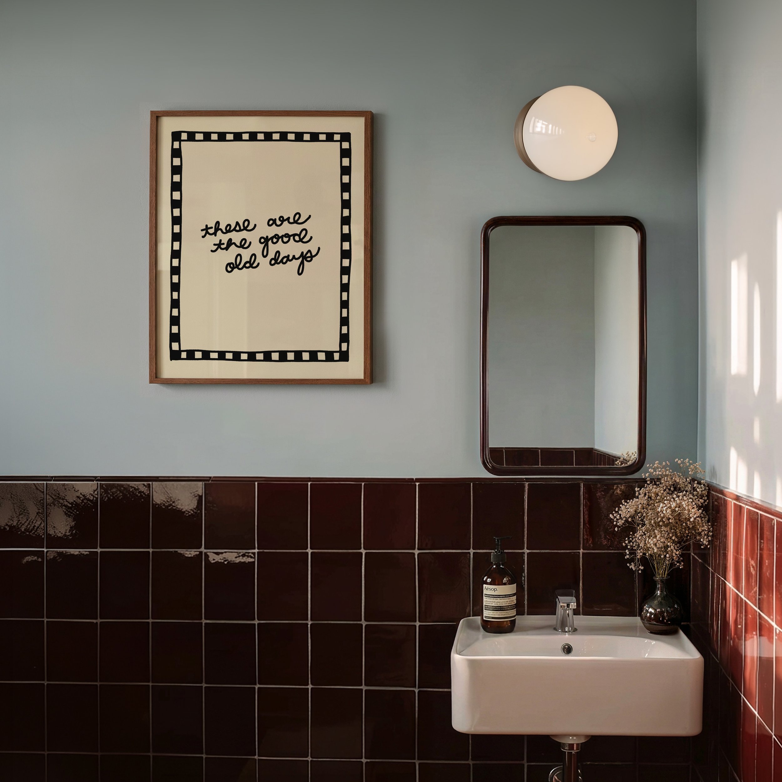 A bathroom wall with a framed poster reading 'those are the good old days', a white circular wall light, a mirror, a small sink with a soap dispenser, a faucet, and decorative dried flowers.