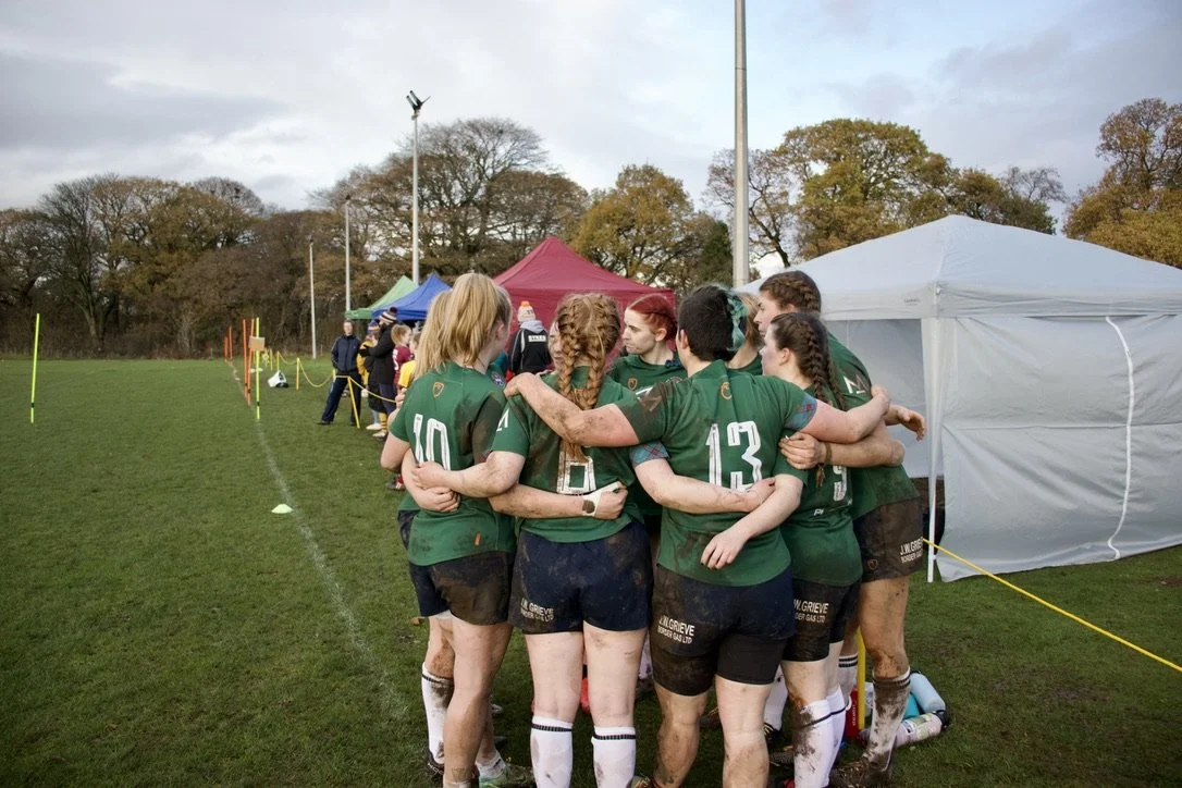 A women's rugby team huddled together on the field during a game, wearing green uniforms with numbers, some with mud on their legs, in a sports field with tents and trees in the background.