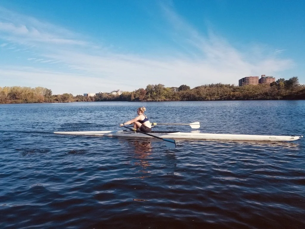 A person rowing a boat on a calm river under a clear blue sky with trees and buildings in the background.