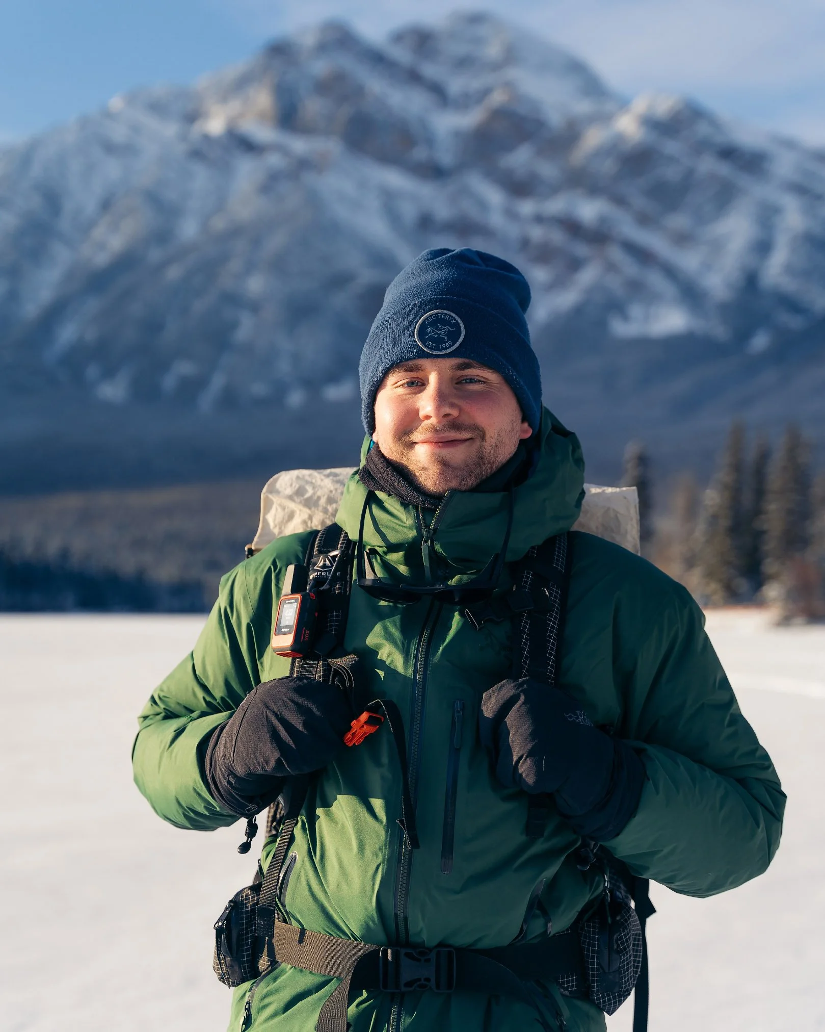 A man in a green and black winter jacket and a dark blue Beanie with a logo stands in a snow-covered landscape with a mountain in the background, smiling at the camera.