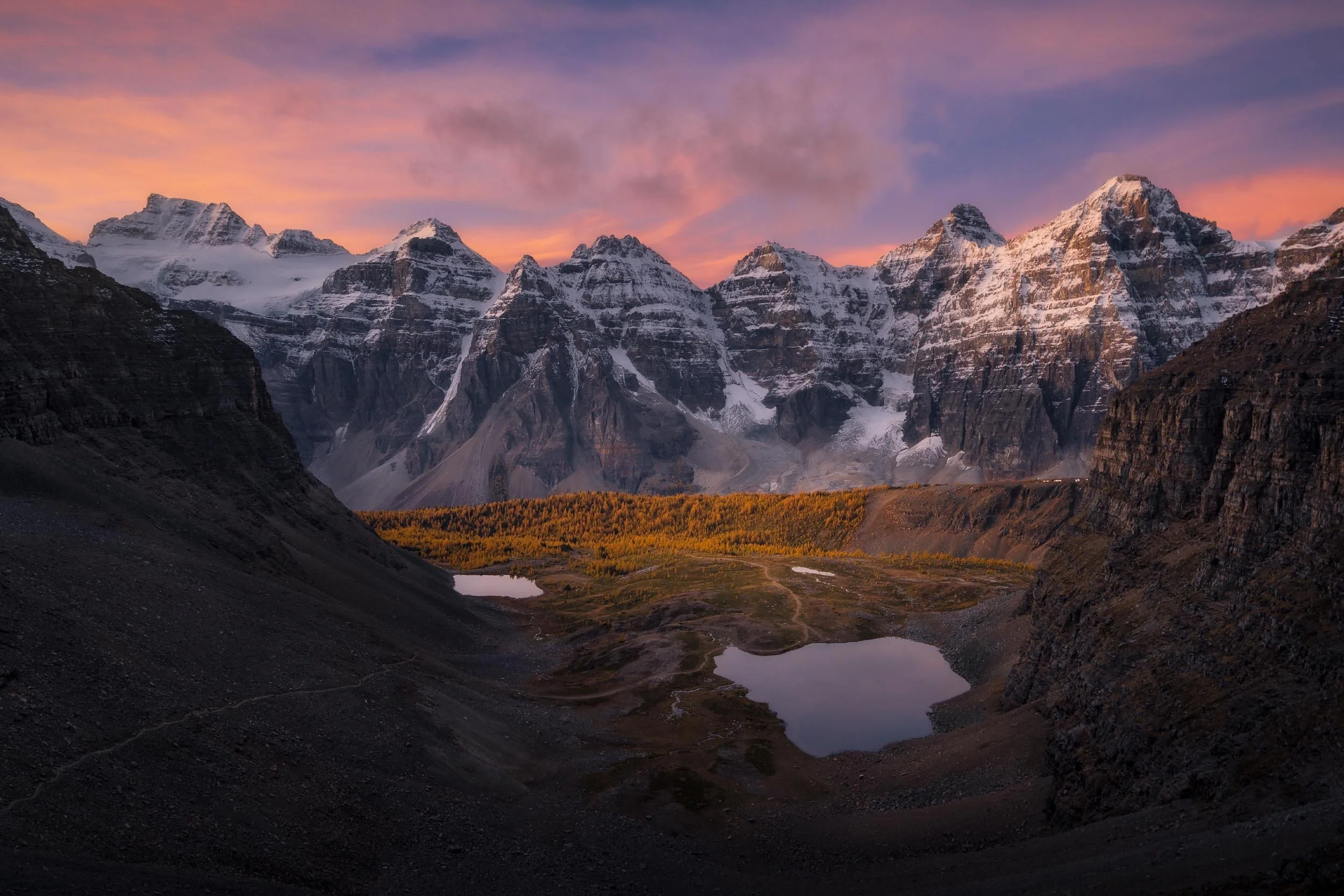 Snow-capped mountains during sunset with pink and orange sky, a valley with a small lake, and rugged terrain in the foreground.