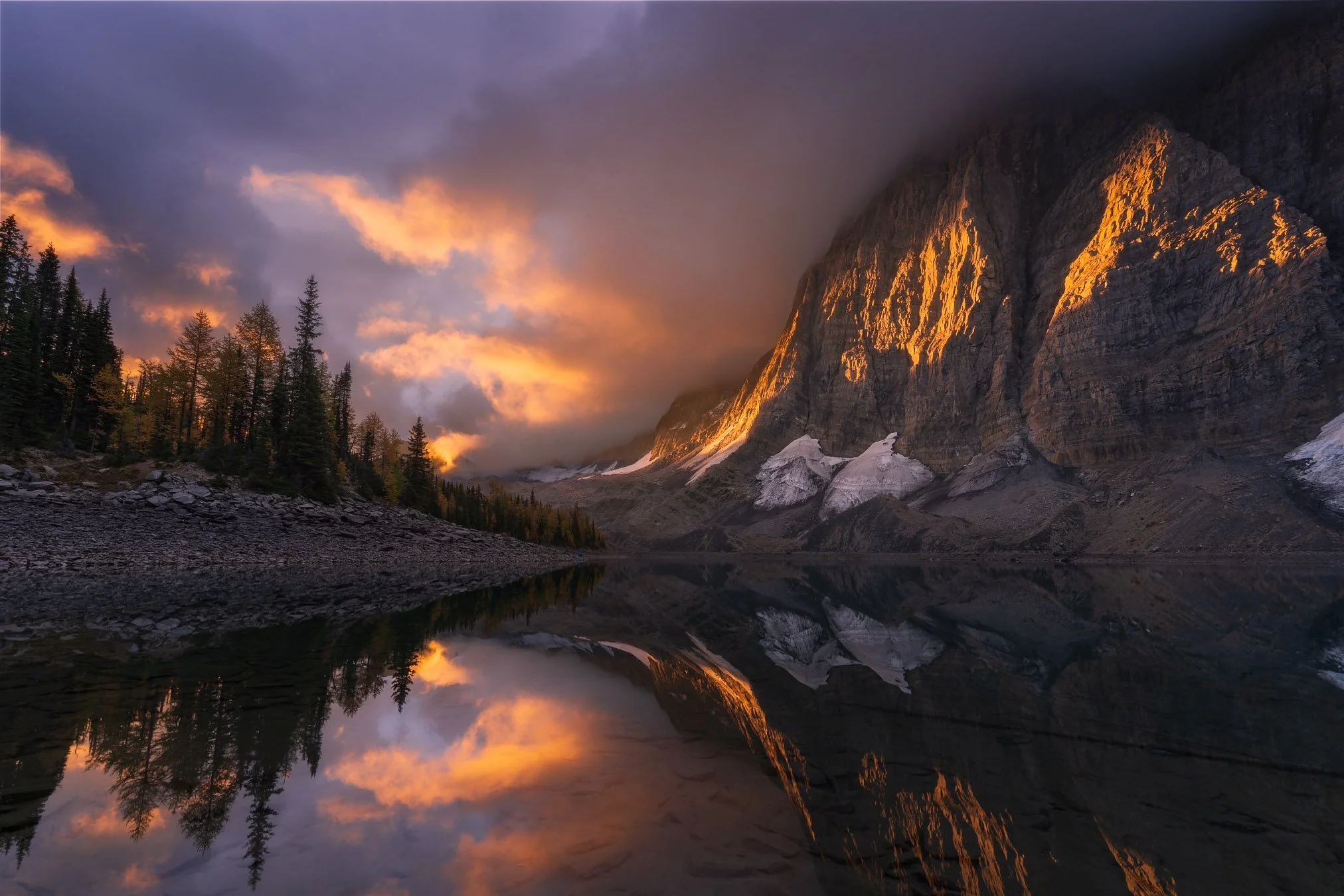 Sunset over a mountain with reflection in a lake, trees on the left side, dark clouds above, and snow patches on the mountain.