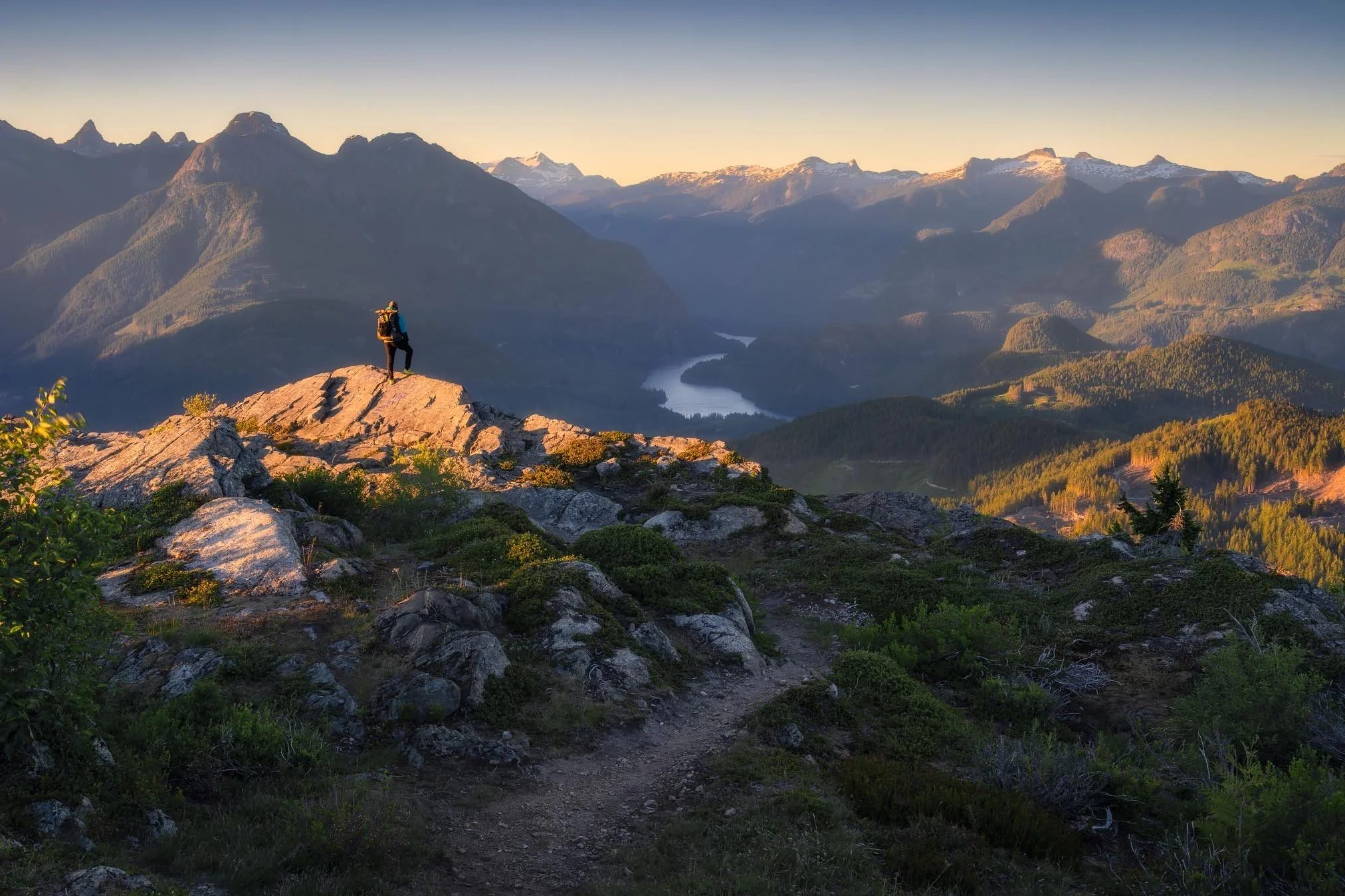 A hiker wearing a backpack standing on a rocky outcrop, overlooking a valley with a river and distant snow-capped mountains at sunset.
