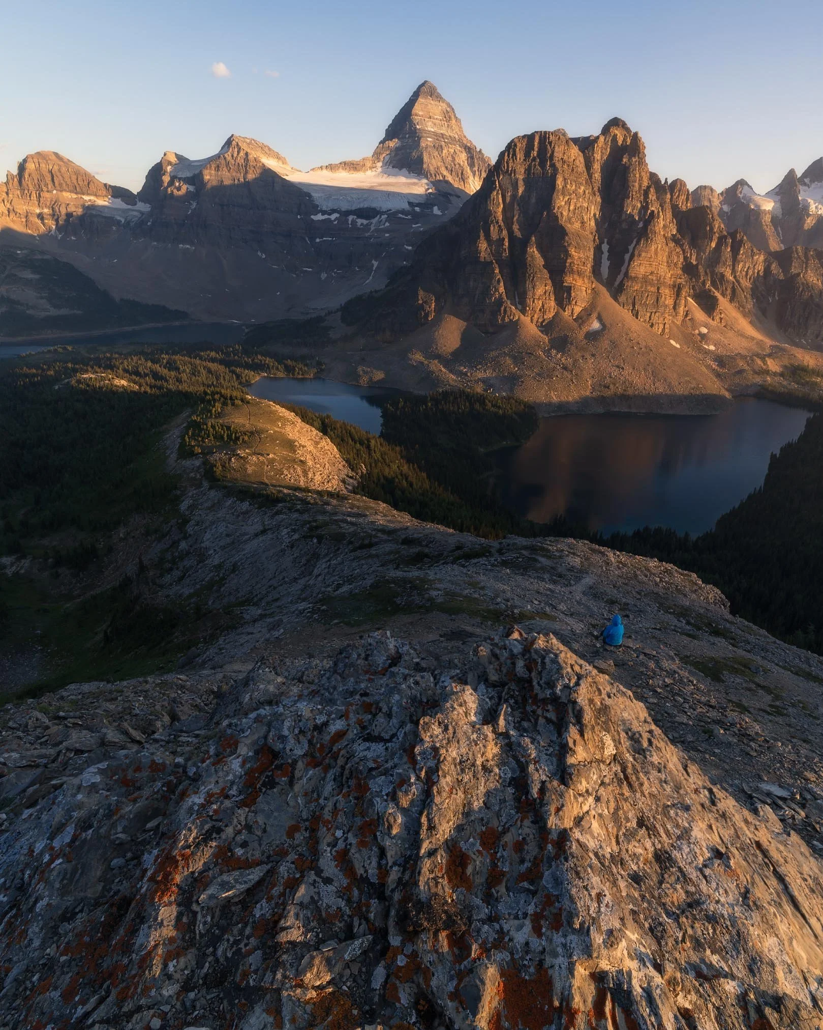 Scenic view of tall, rocky mountains with snow patches, a lake at the base, and a person sitting on the rocky foreground during sunset or sunrise.