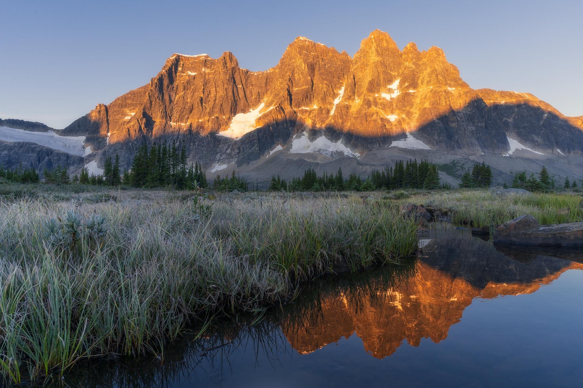 Sunlit mountain range with snow patches, next to a calm river reflecting the mountains and the clear sky.