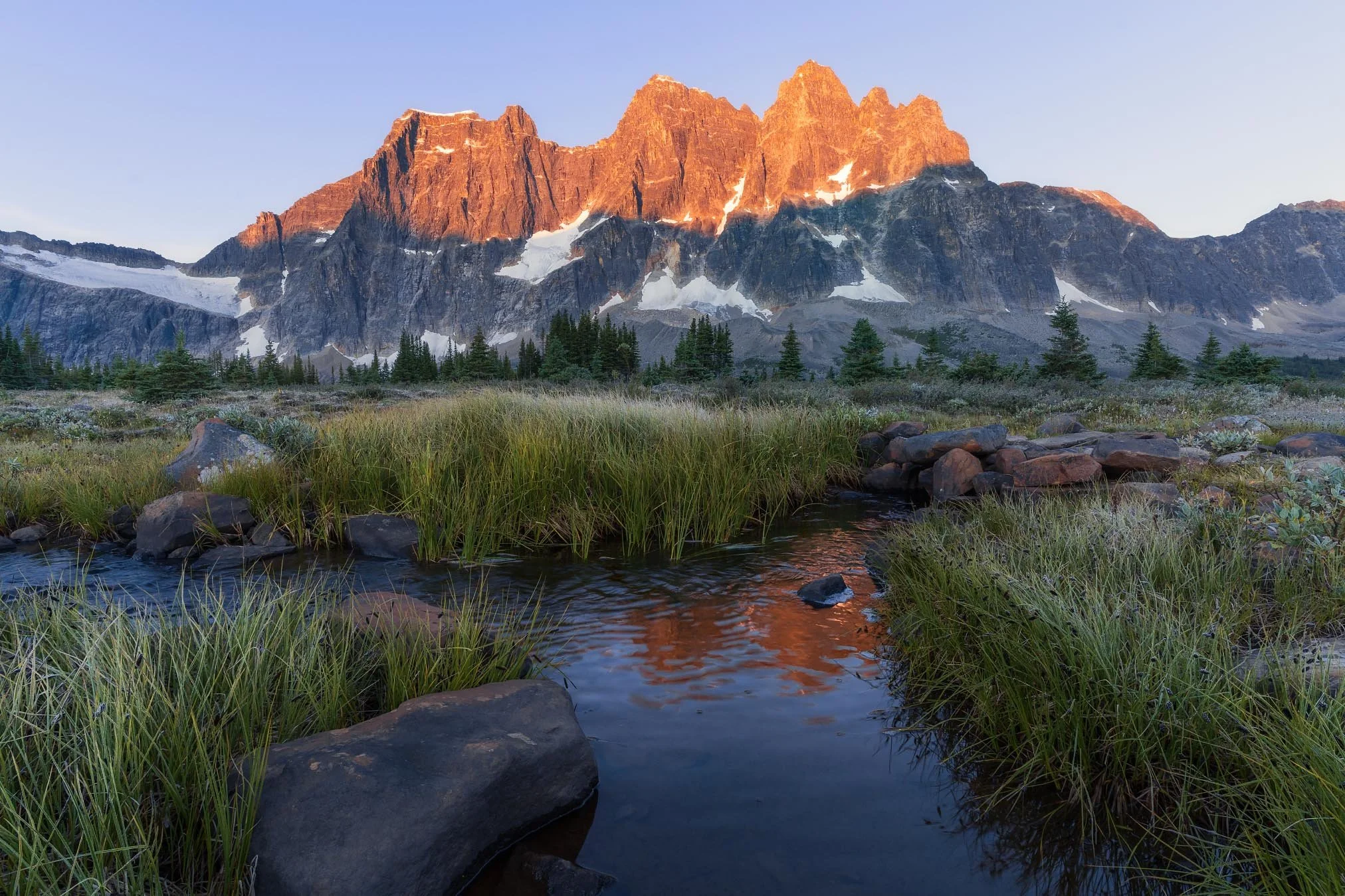 Sunset lighting on a mountain range with snow patches, green trees, and a small stream in the foreground.