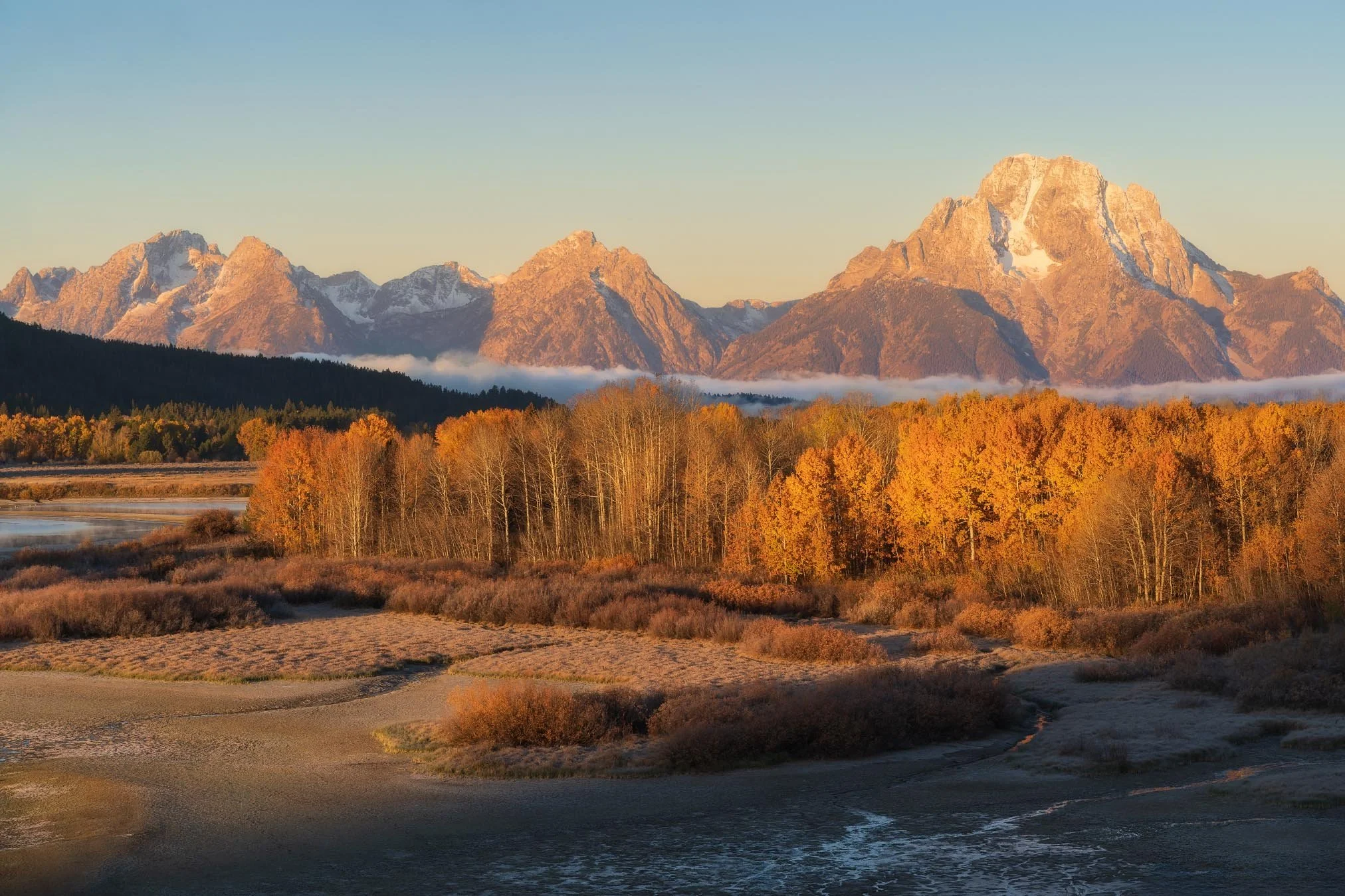 Landscape of majestic snow-capped mountains during sunset, with a forest in fall colors and a winding dirt path in the foreground.