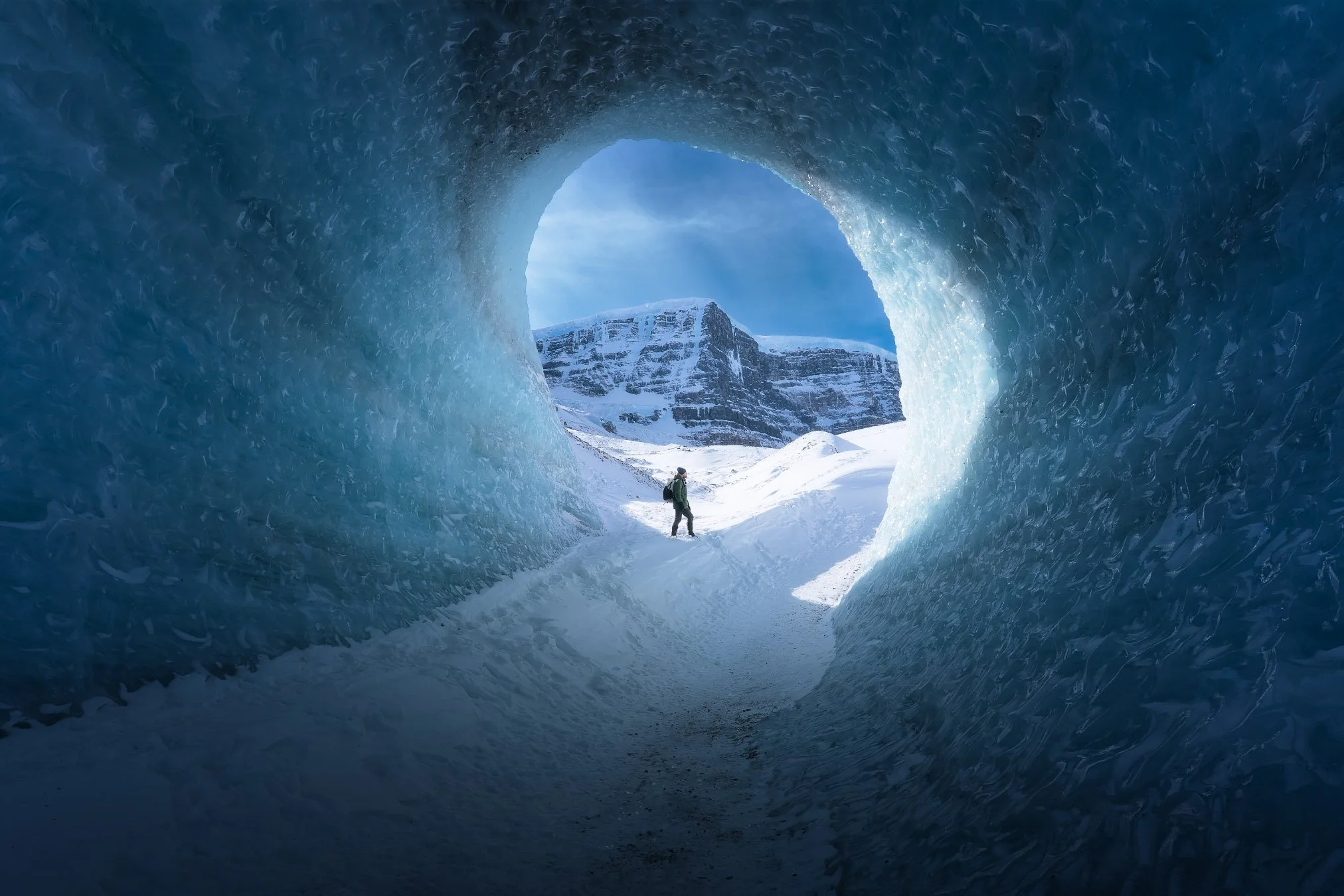 View from inside an ice cave looking out at snow-covered mountains and a person walking on snow, with blue ice walls surrounding the entrance.