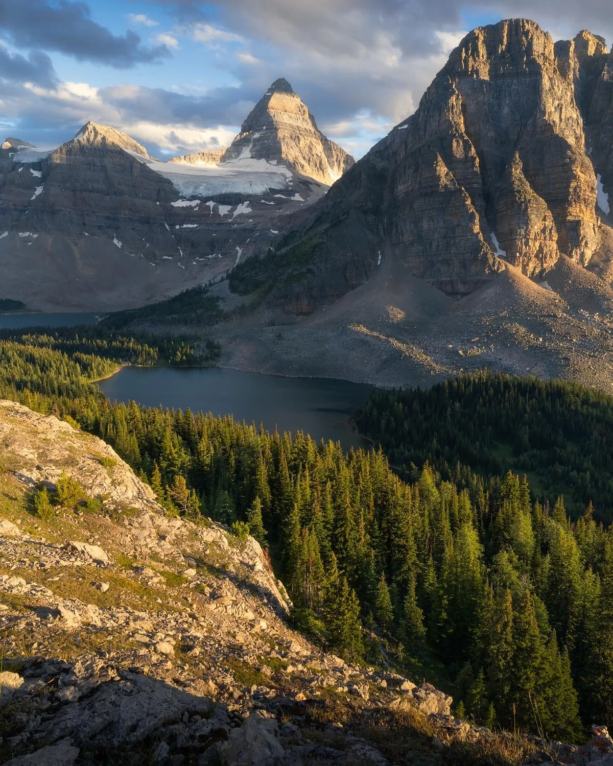 A mountain landscape with three prominent peaks, snow patches, a lake at the base, dense forest, and partly cloudy sky, illuminated by golden sunlight.