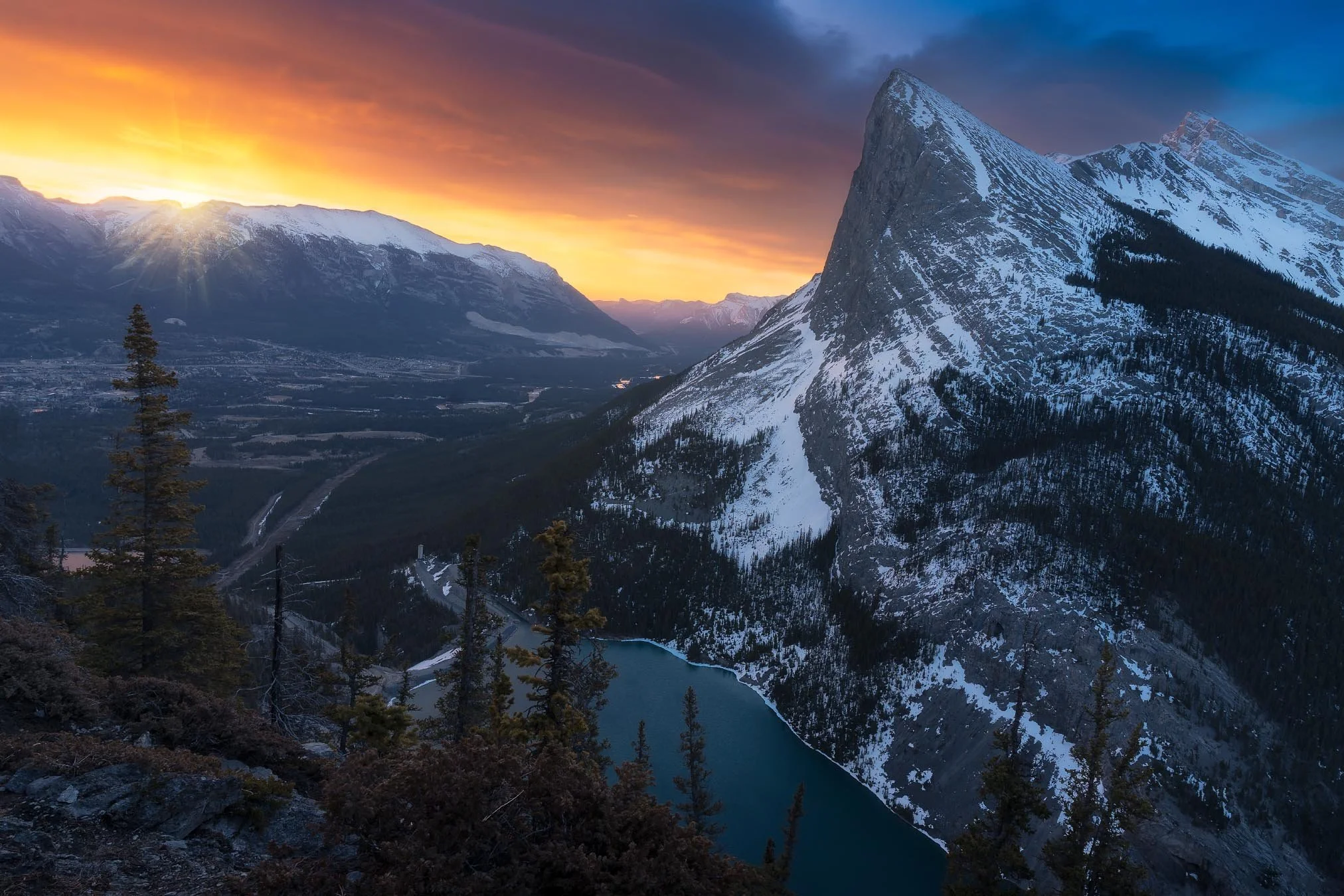 Snow-capped mountain range with a prominent, steep, pointed peak illuminated by sunset, with a winding river and pine trees in the foreground, and a colorful sky with orange and purple hues.