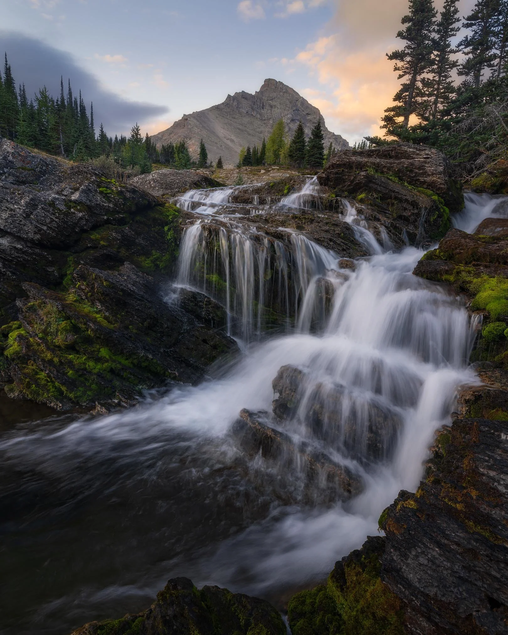 A scenic mountain landscape with a waterfall flowing over rocks in the foreground, tall trees along the sides, and a mountain peak in the background under a partly cloudy sky at sunset or sunrise.