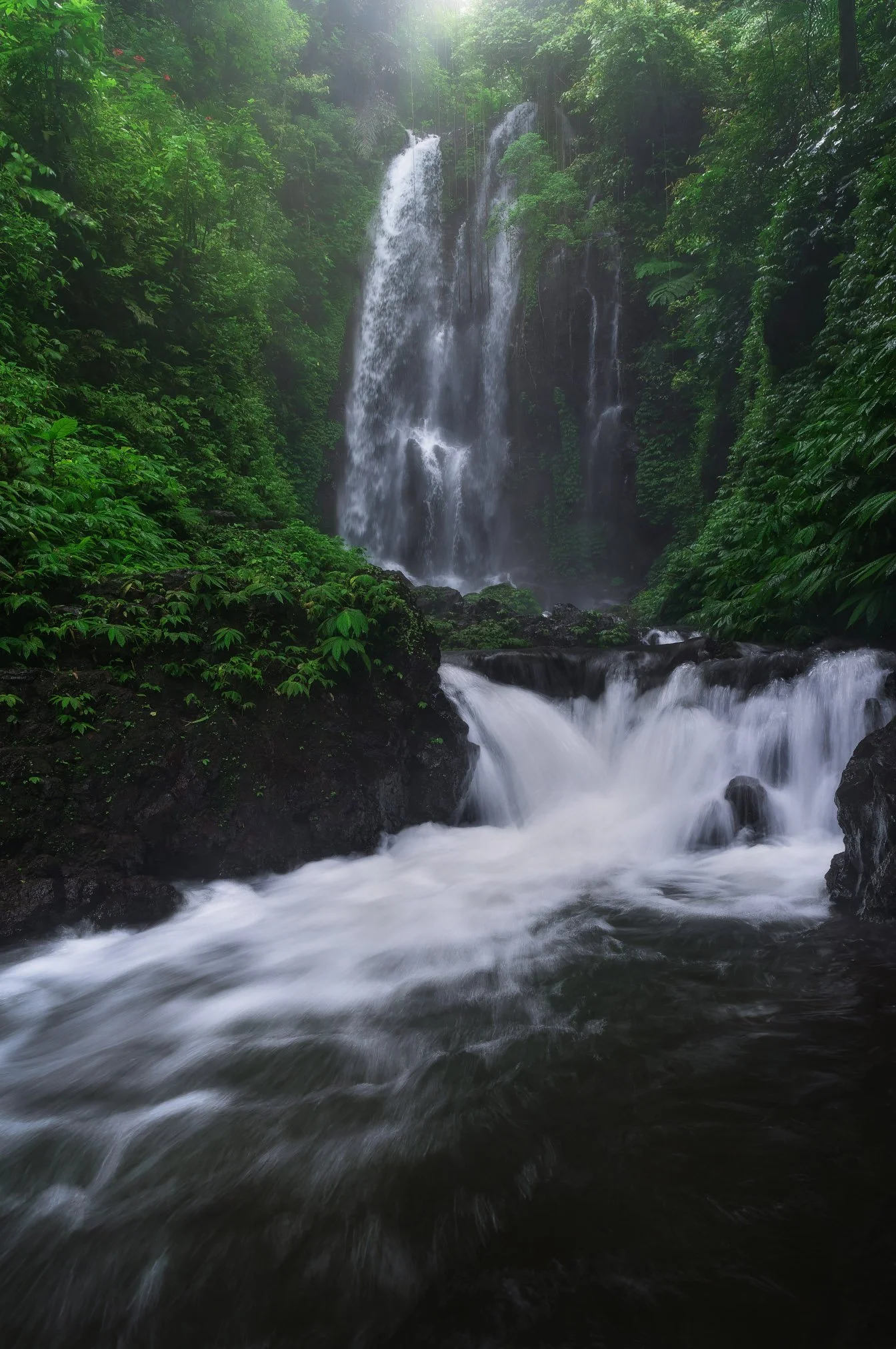A multi-tiered waterfall flowing down a lush, green forested cliffside with water rushing over rocks in the foreground.