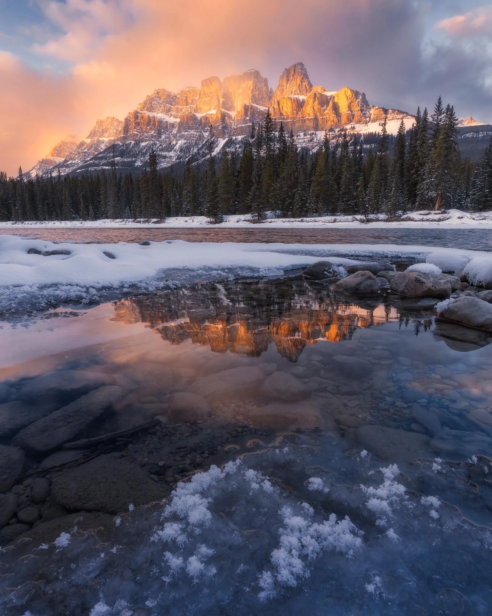 Snow-covered shoreline with rocks in the foreground, dense evergreen trees in the middle ground, and a mountain range illuminated by sunset in the background.
